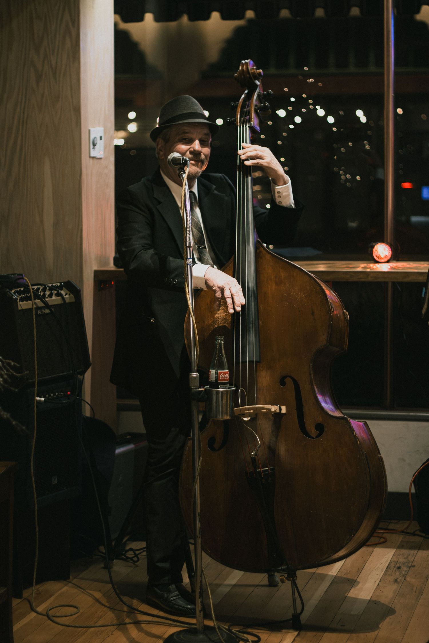 A jazz musician in a suit and fedora plays double bass at a dimly lit venue.