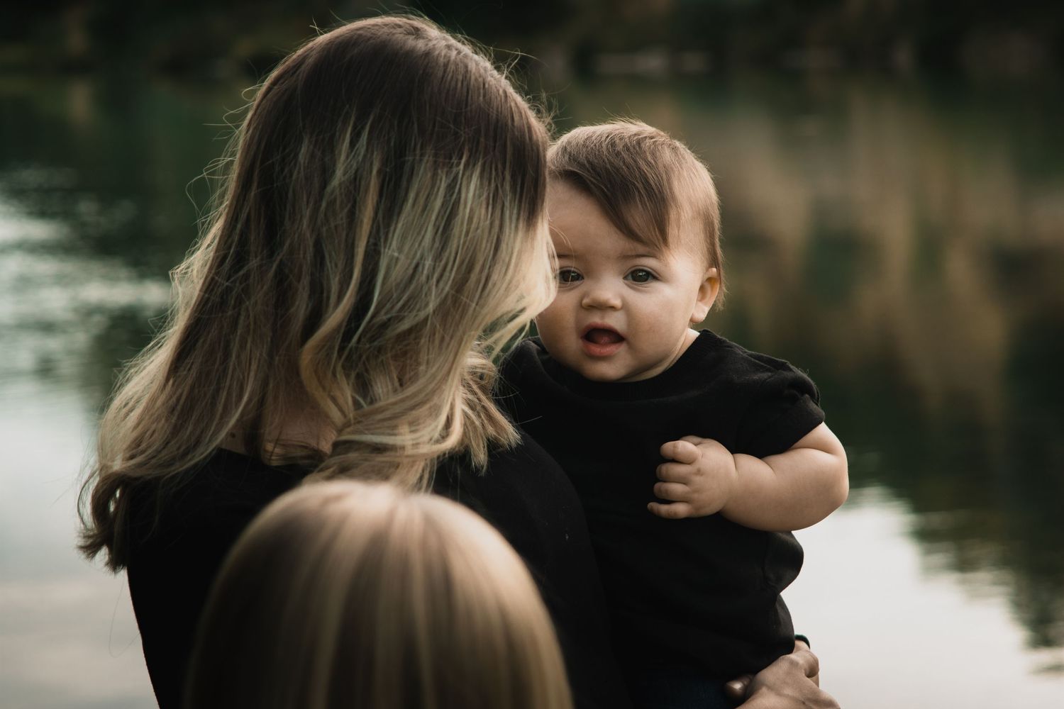 A tender moment between parent and child in black clothing by a tranquil lake during golden hour.