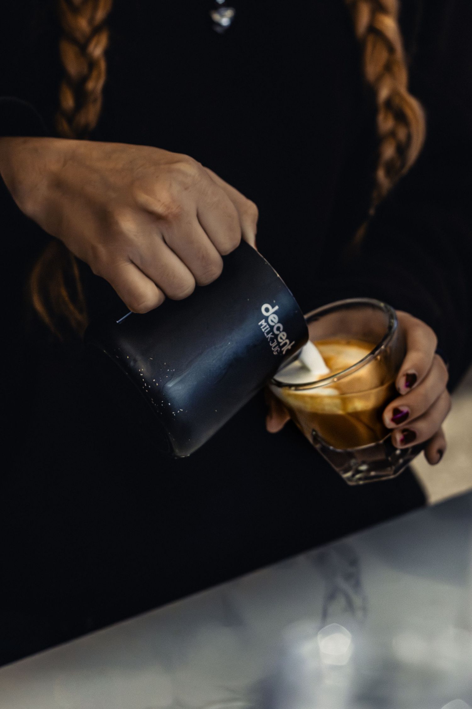 Hands pouring steamed milk into a glass of iced coffee.
