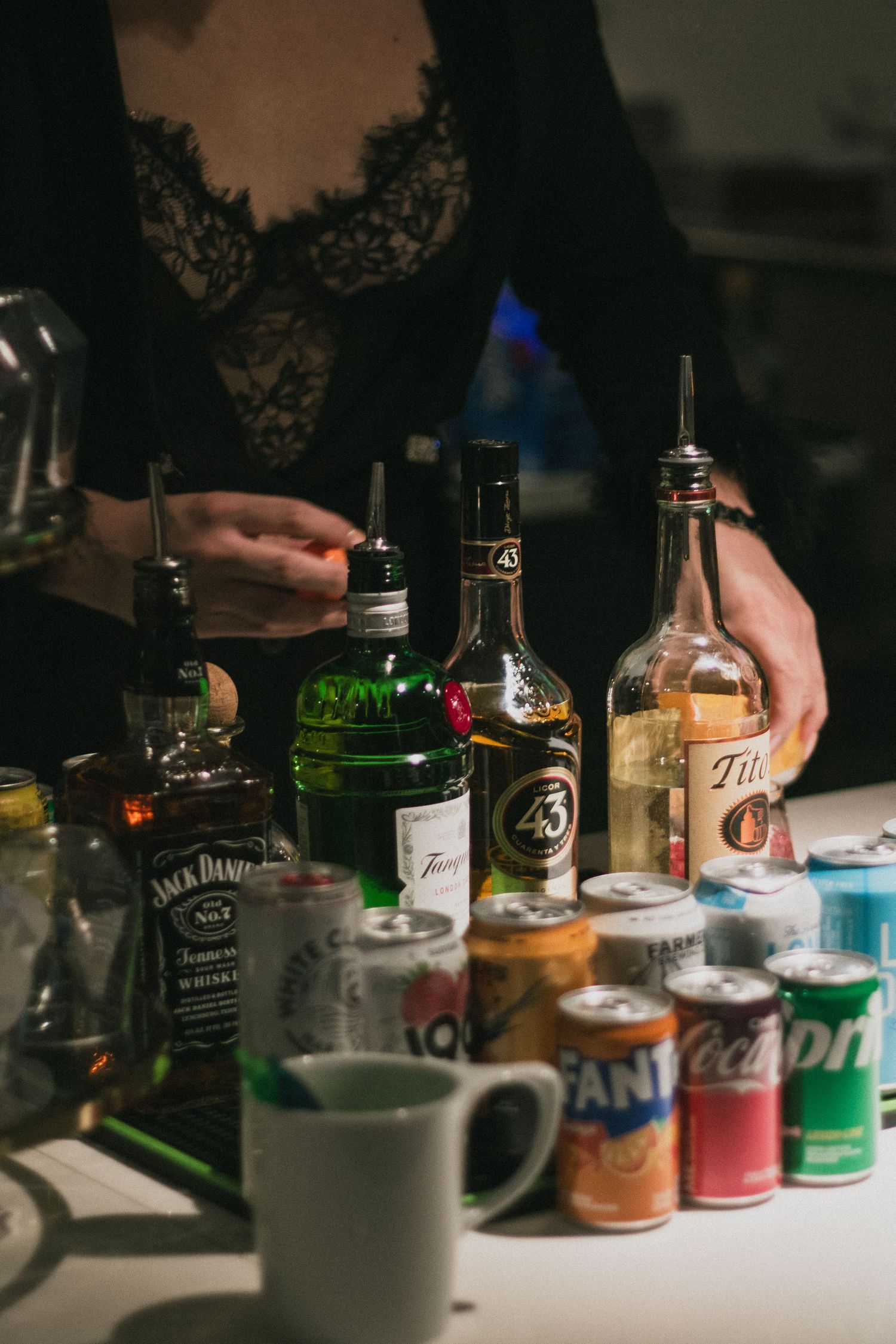 A bartender arranges bottles of Jack Daniels and liqueur with soda cans on a bar.