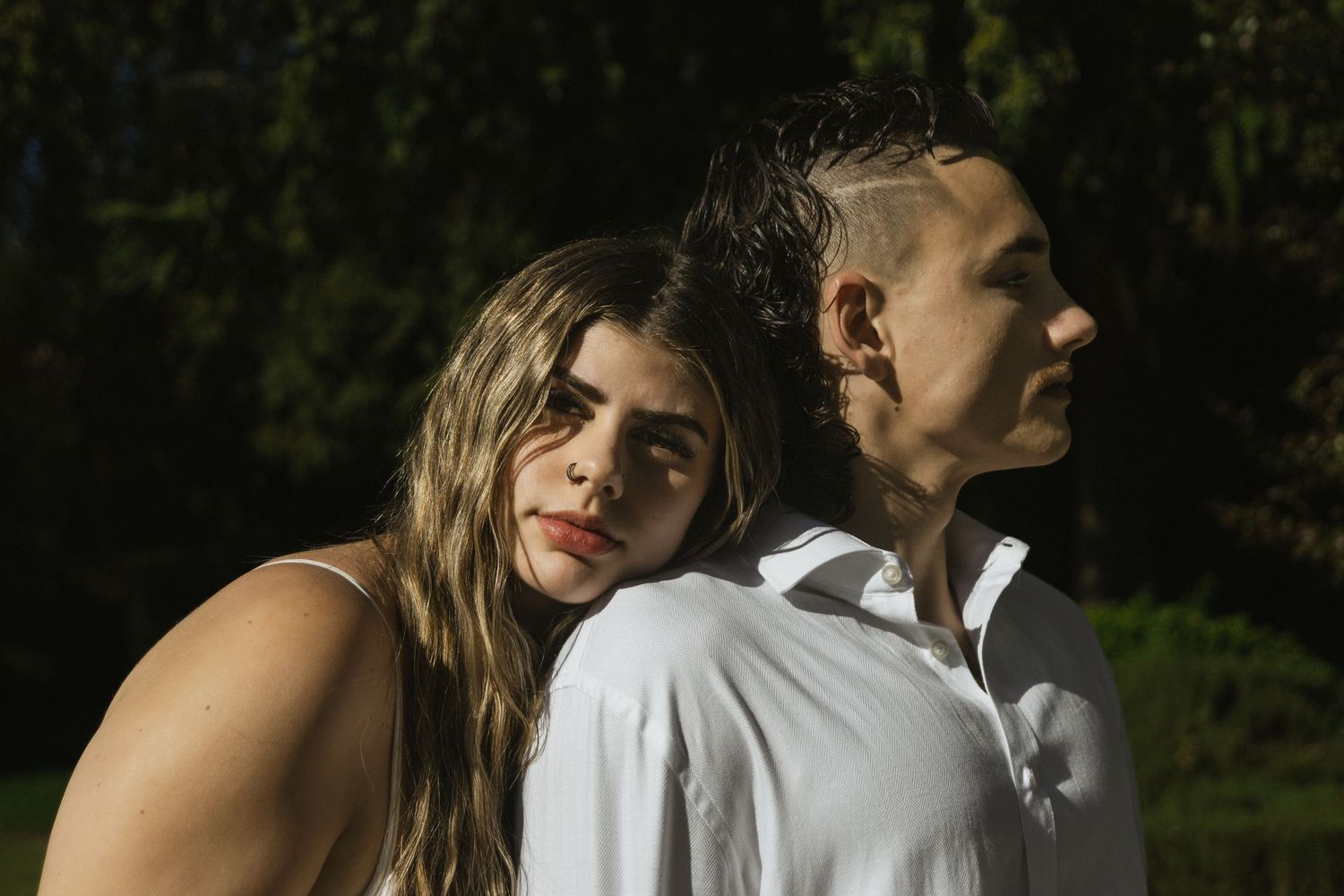 Couple in white clothing standing together in dappled sunlight outdoors.