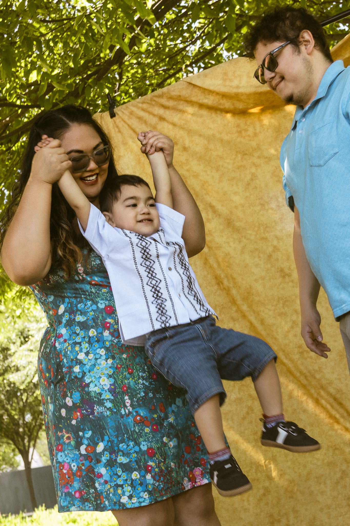 Family of three outdoors, mother in floral dress and father in blue shirt lifting smiling toddler boy near yellow backdrop.