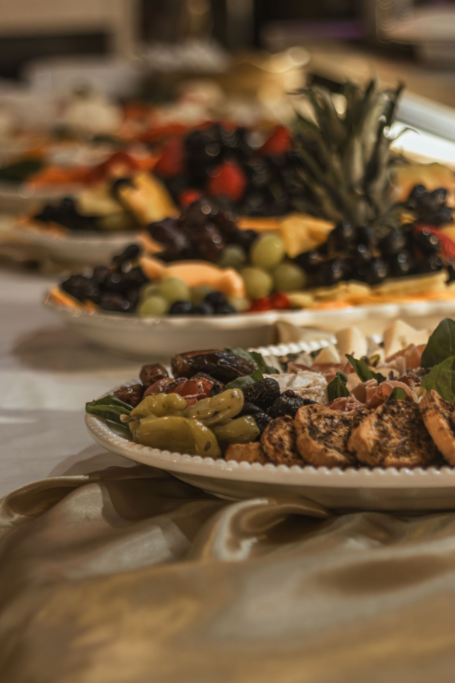Elegant catering spread with plates of olives, grilled meats, fresh fruits, and a pineapple centerpiece on a white tablecloth.