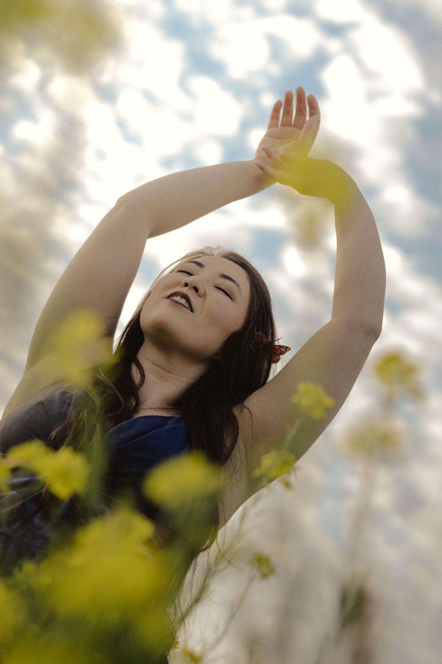 A person raises their arms joyfully against a bright cloudy sky with yellow flowers in the foreground.