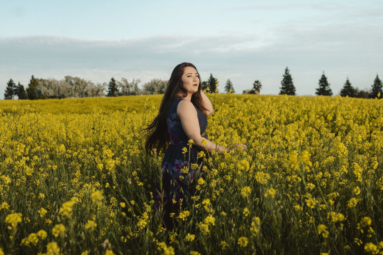 Figure in dark dress poses elegantly among blooming yellow canola flowers at sunset.