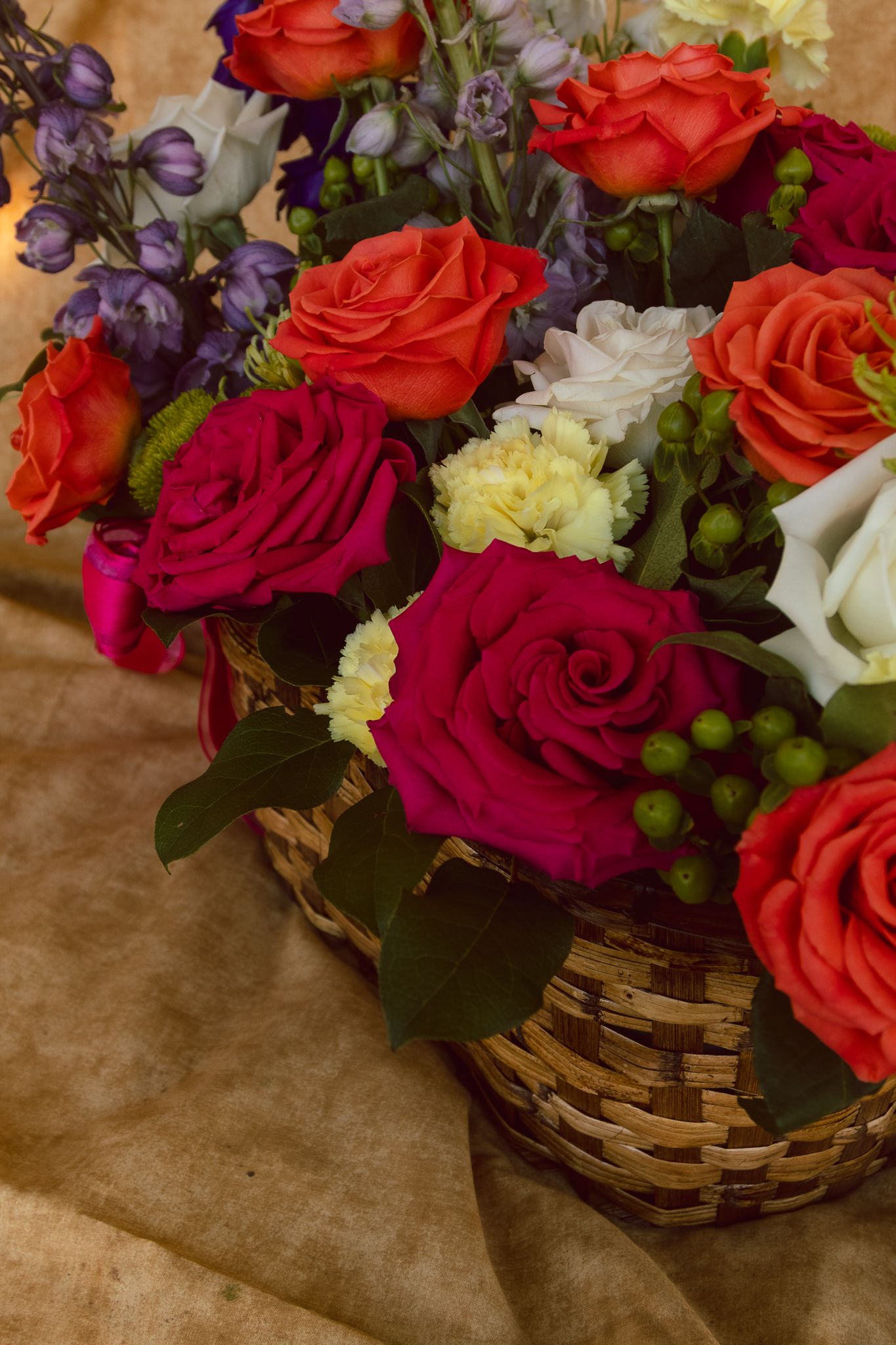 Colorful floral arrangement in wicker basket featuring red, orange roses, purple flowers, and green berries on brown paper.