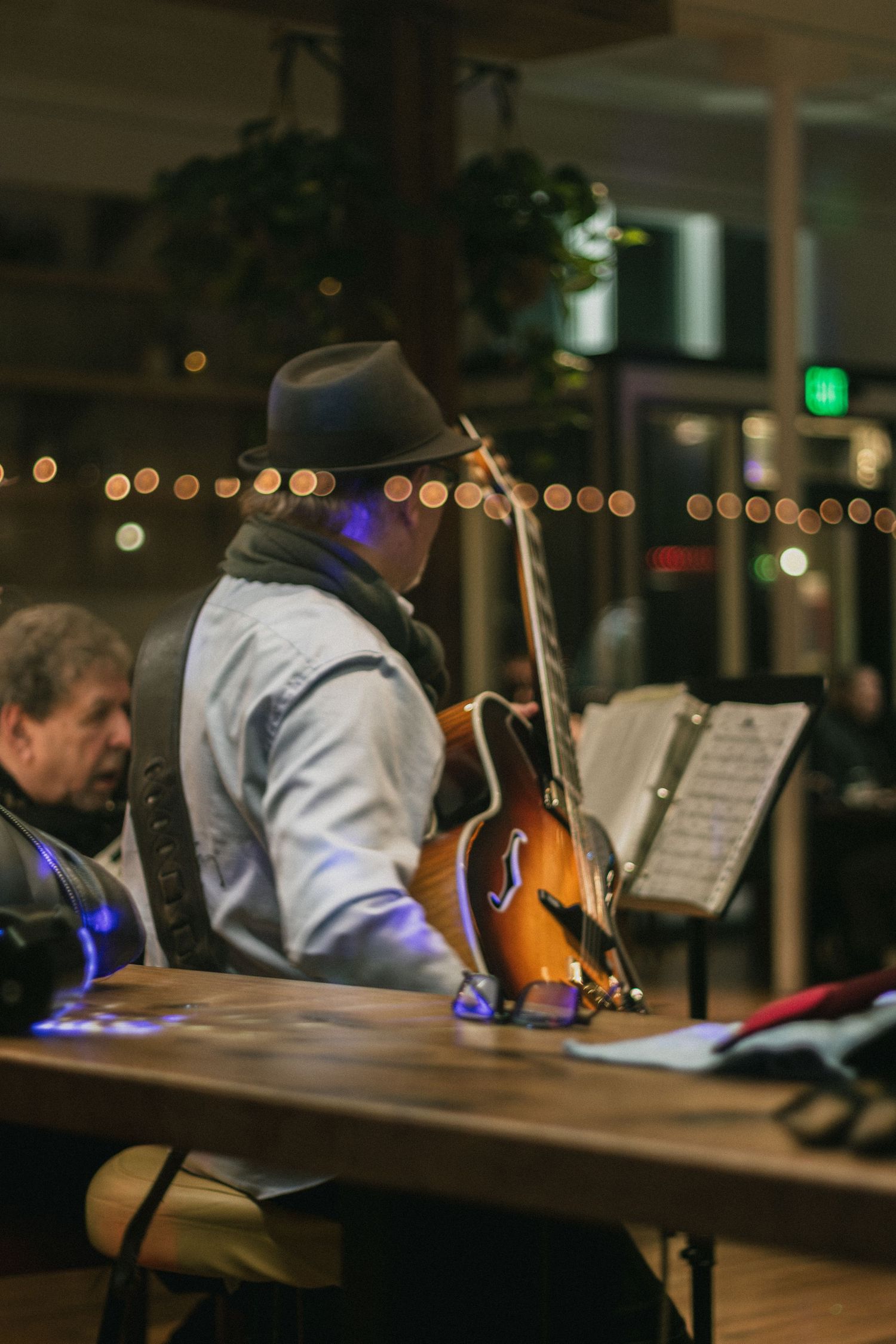 A musician in a hat plays guitar at a dimly lit indoor venue with string lights in the background.