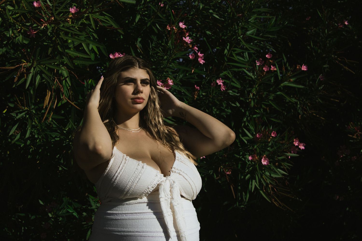 A woman in white poses against flowering bushes in a garden setting.