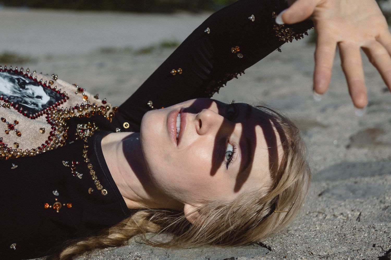 Blonde woman lying on sand with eyes closed, wearing embroidered black top, with dramatic shadows across her face.