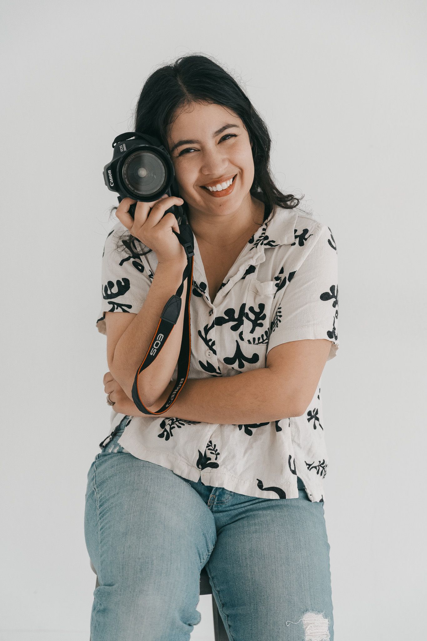 Photographer in blue jeans and floral shirt poses playfully with professional camera against white background.