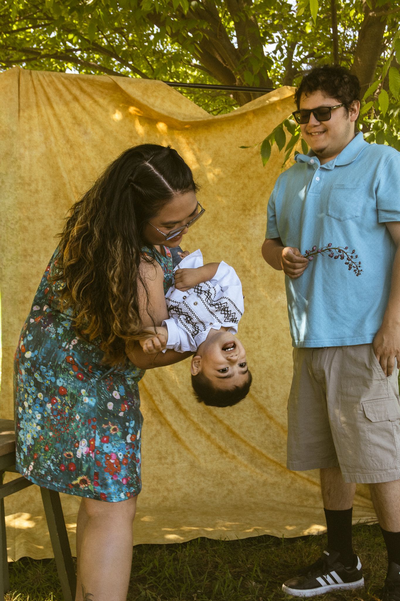A woman in a floral dress playfully holds a baby upside down while a man in a blue polo smiles outdoors.
