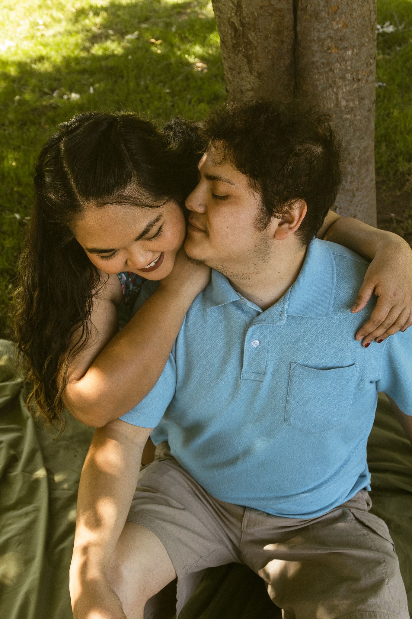 A young couple sharing a tender moment outdoors, sitting together near a tree on green grass.