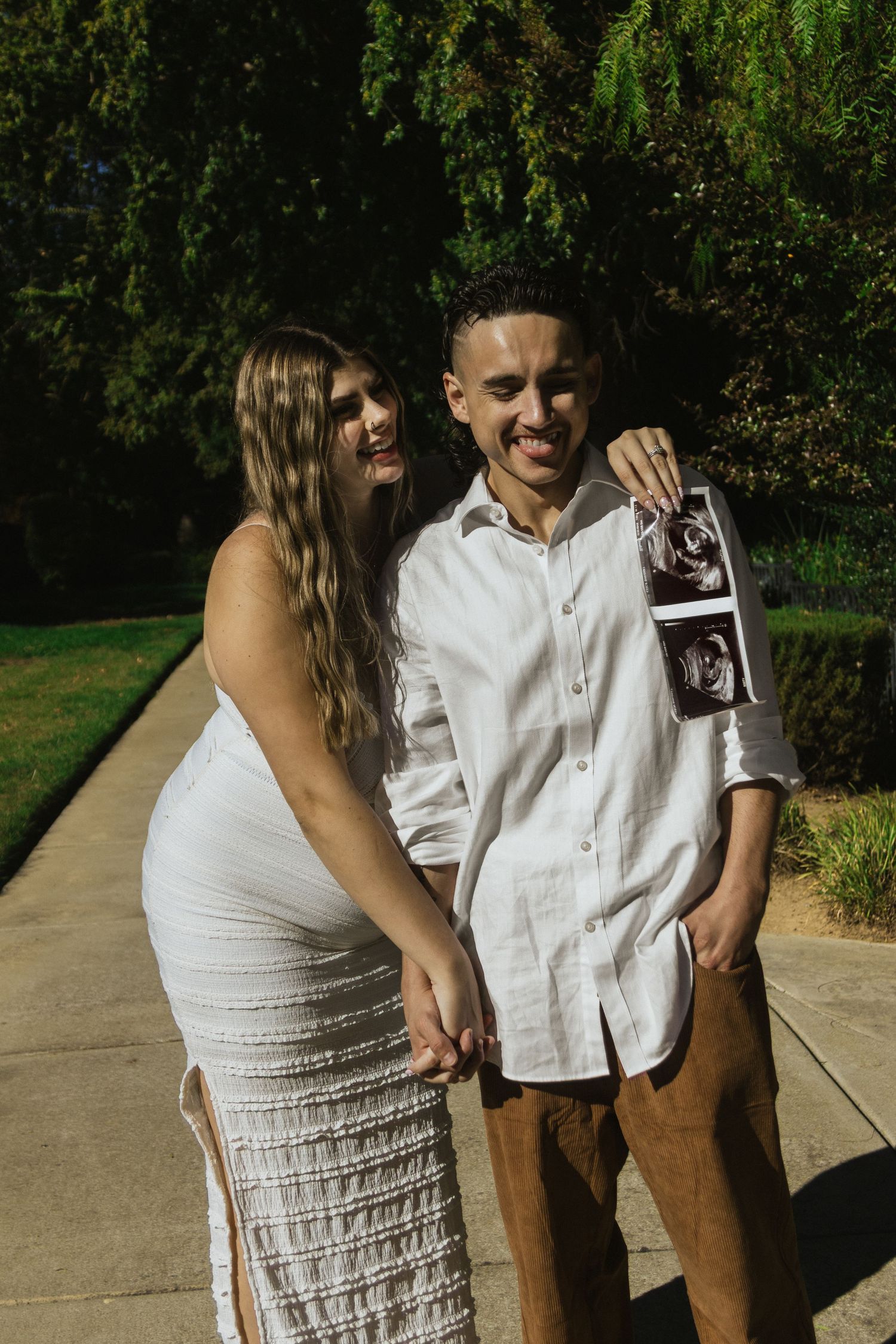 Couple in white and tan outfits sharing a joyful moment on a sunny pathway.