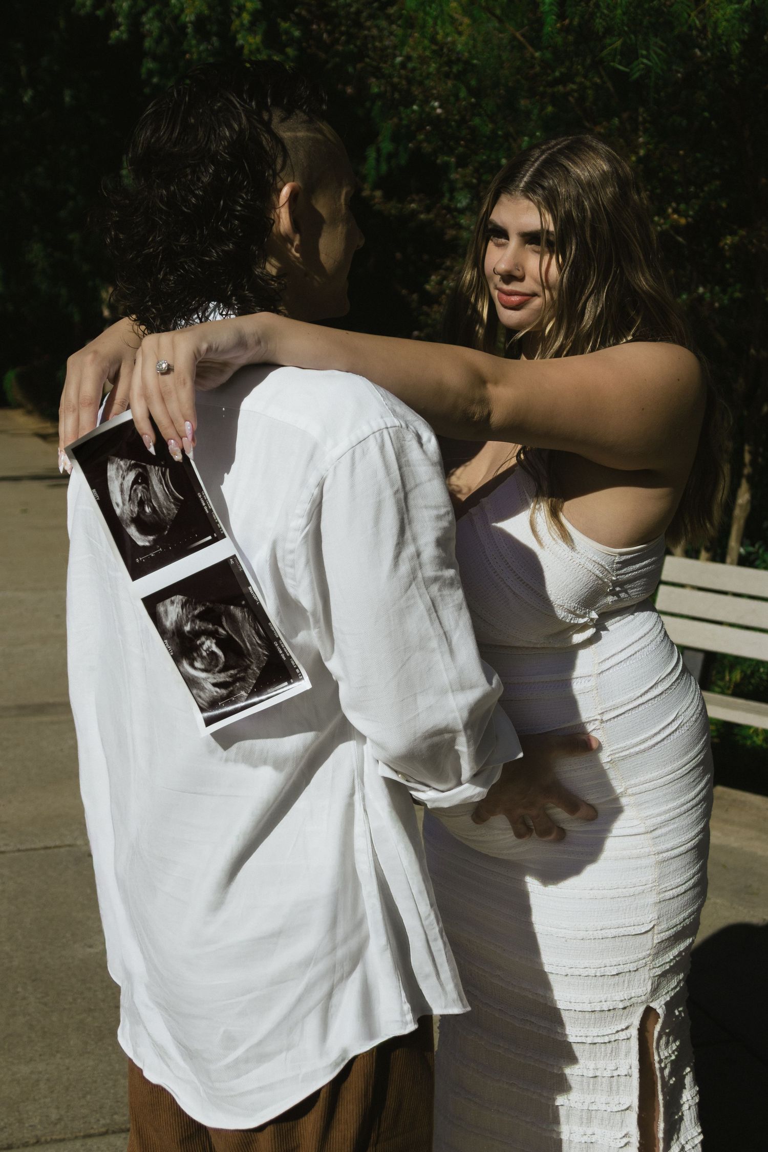 A couple sharing an embrace while holding ultrasound photos outdoors.