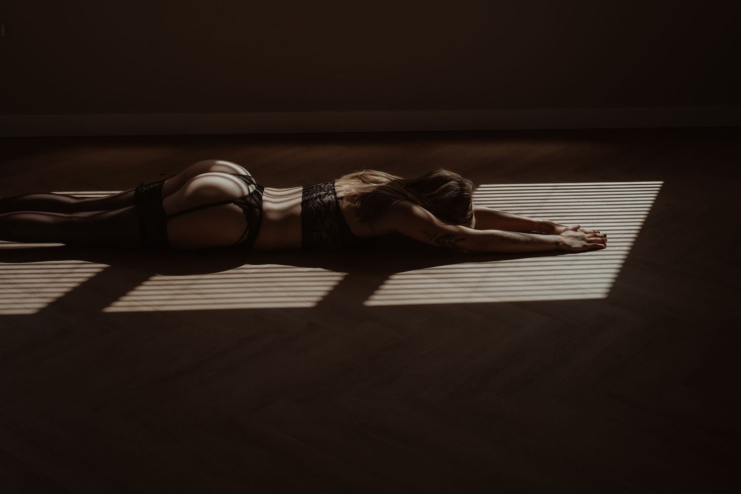 Woman in dark lingerie lying on floor in dramatic shadow and light, artistic boudoir photography.