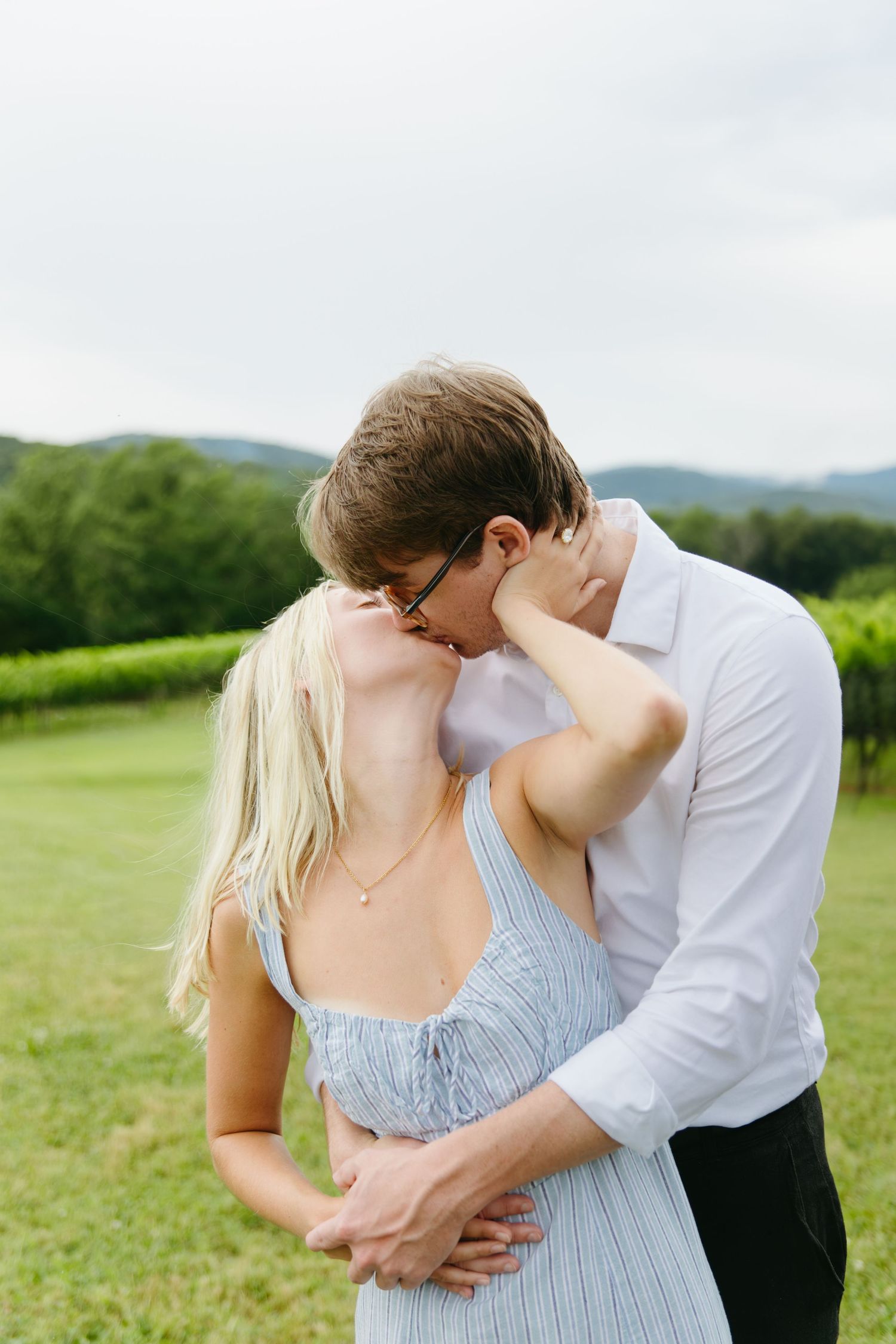 Man in white shirt kisses woman on the cheek from behind as they embrace in front of a green vineyard.