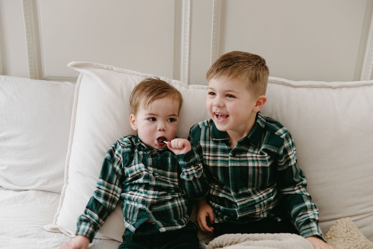 Two young brothers in matching green plaid pajamas sitting together on a white couch.