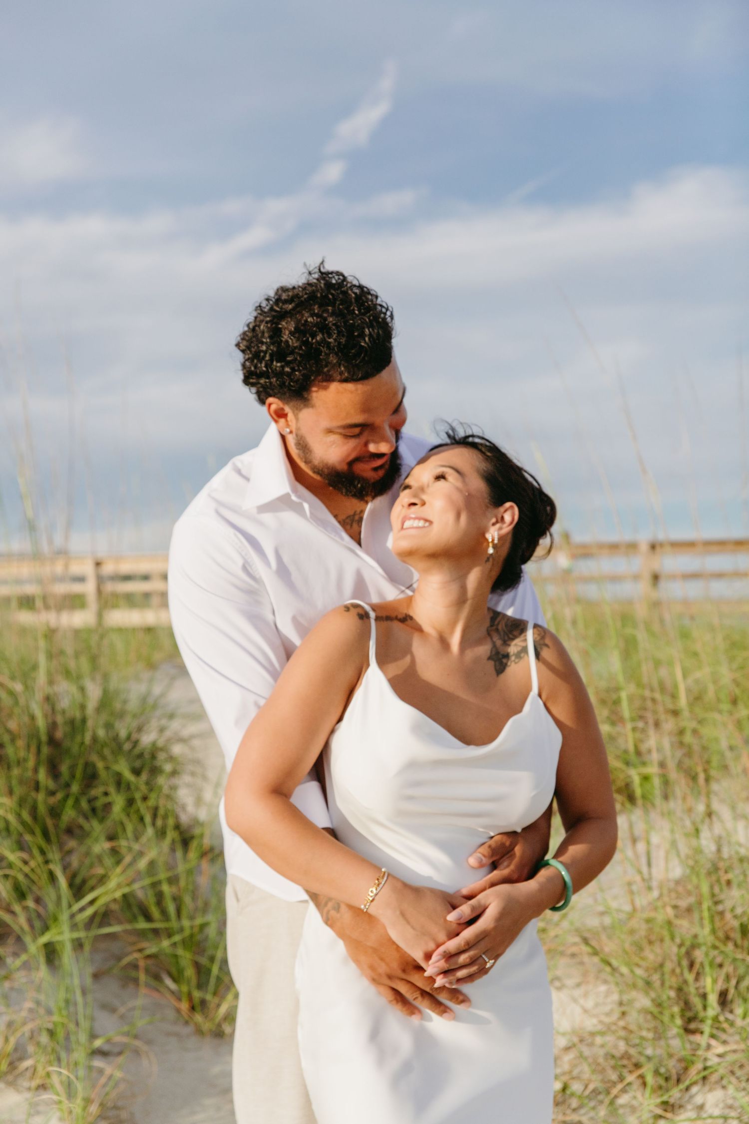 Smiling couple in white holding hands on beach dunes during engagement session.