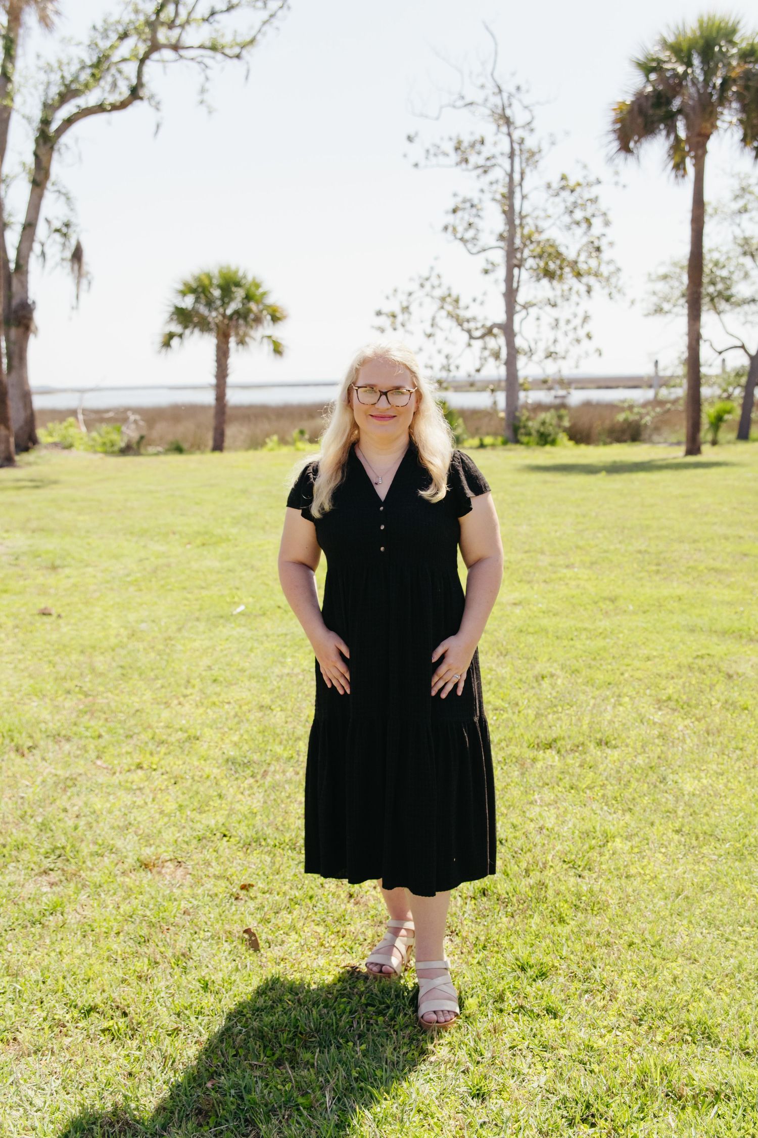 Young woman in black midi dress standing on green lawn with palm trees and ocean in background.