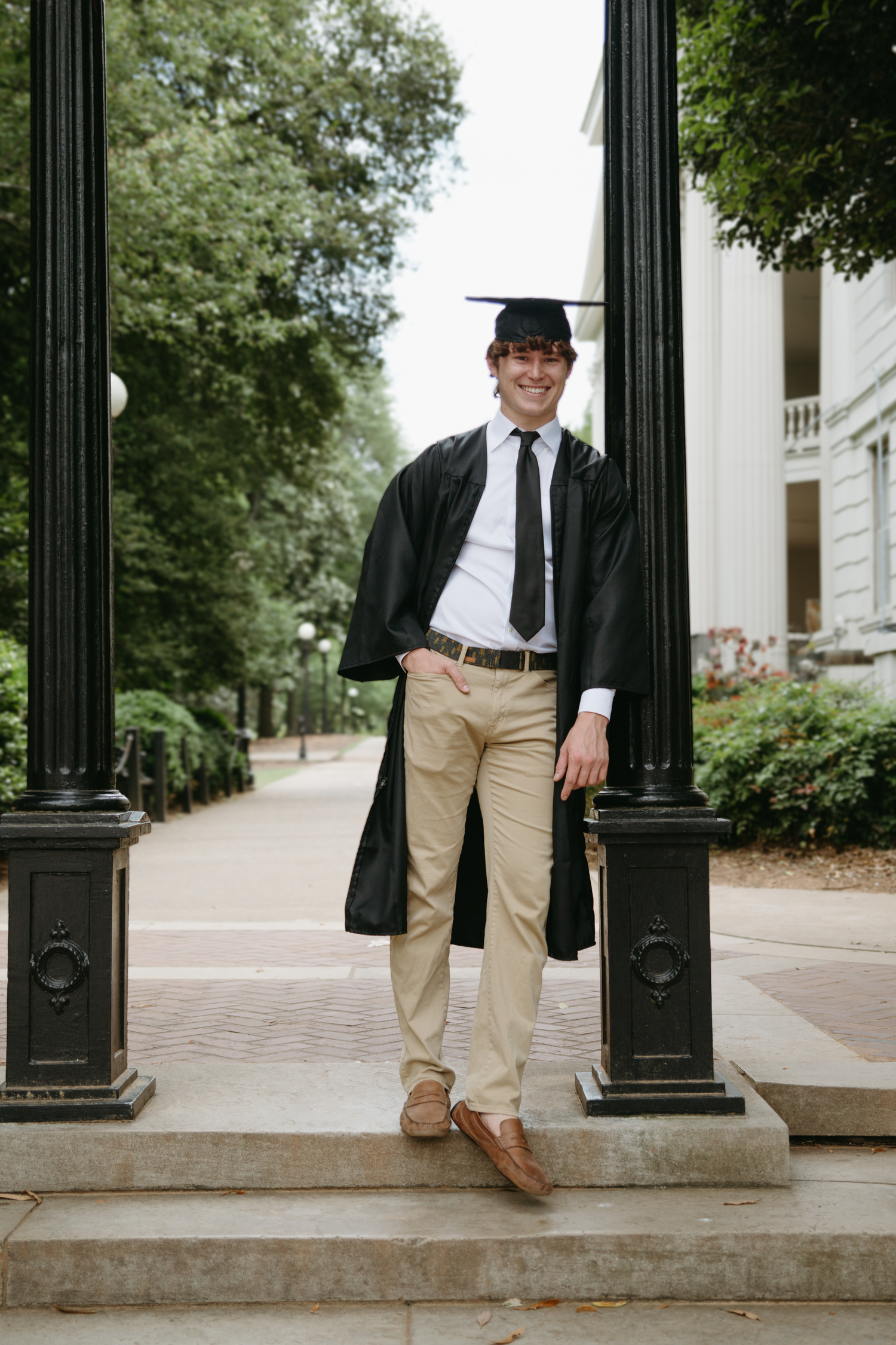 Smiling graduate in cap and gown stands between black pillars on a tree-lined campus walkway.