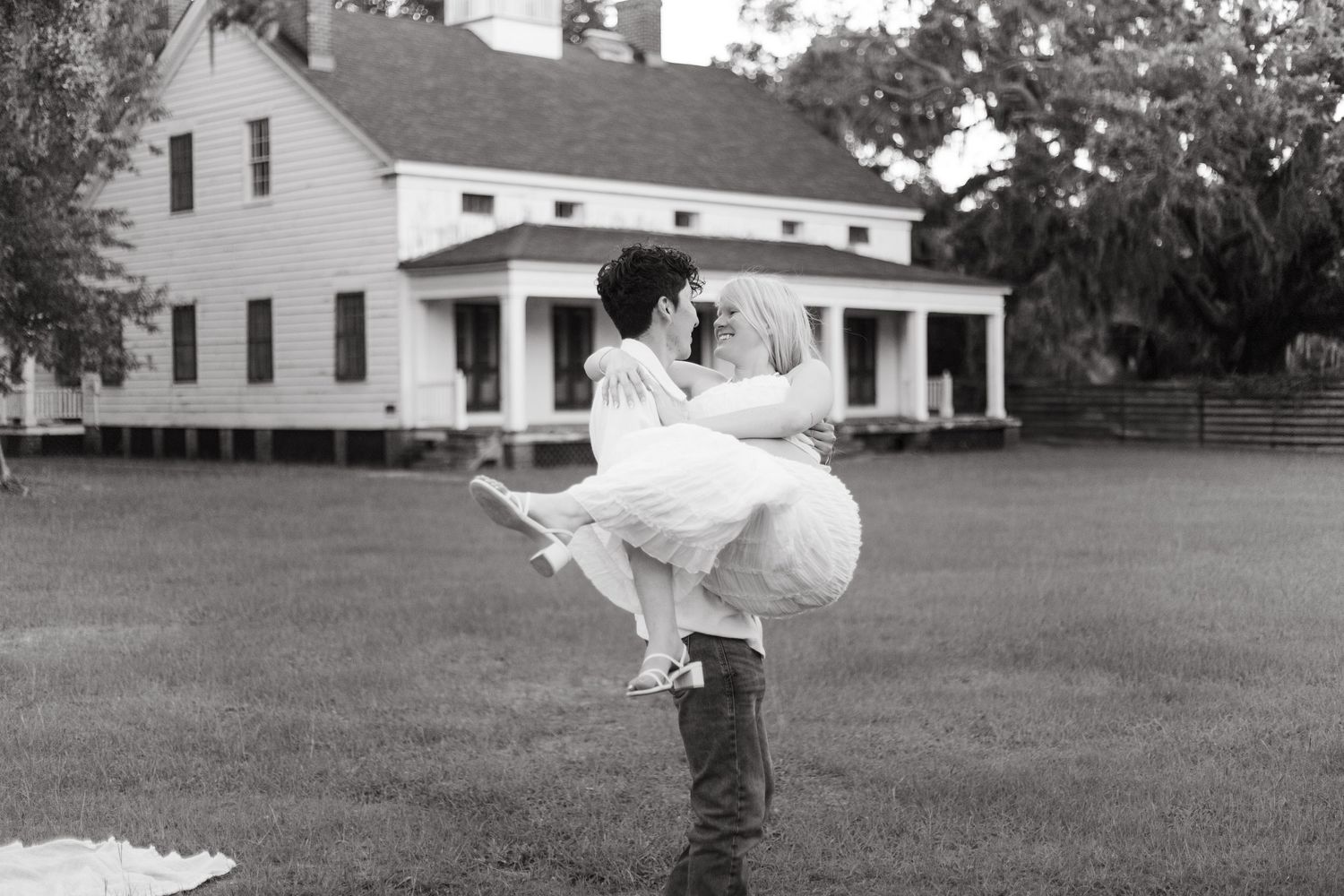 Black and white photo of man lifting pregnant woman in front of a white farmhouse.
