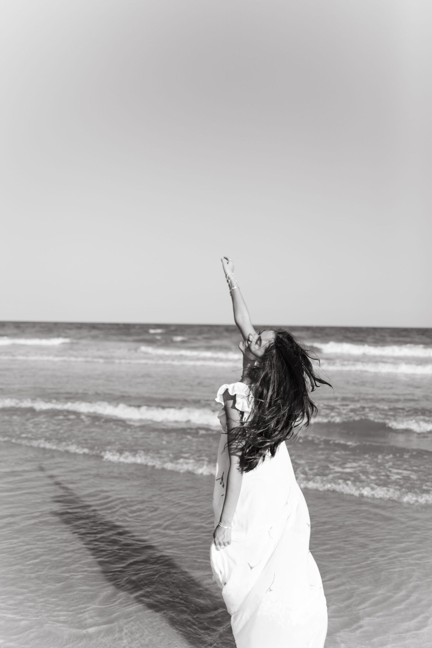 Black and white photo of woman in white dress raising arm on a breezy beach.