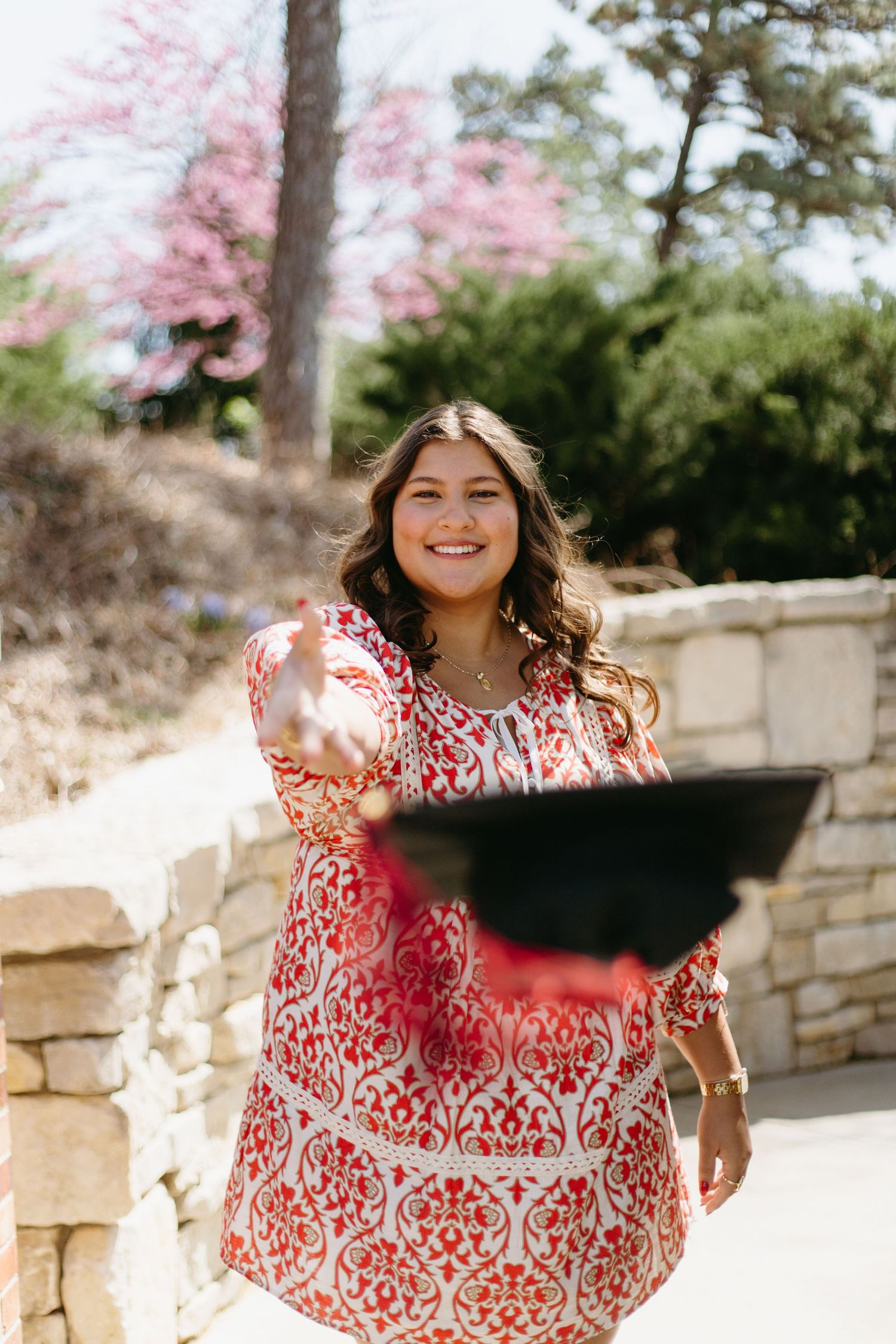 Happy graduate in red patterned dress tossing graduation cap outdoors near blooming pink trees.