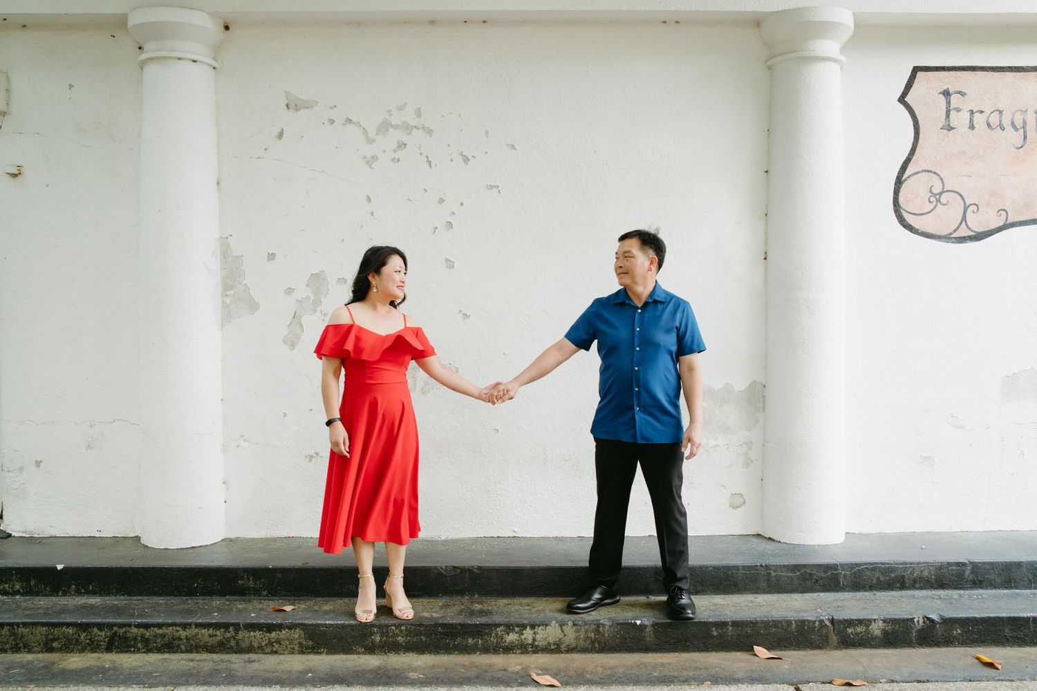 Couple in red dress and blue shirt hold hands on steps in front of a white wall outdoors.