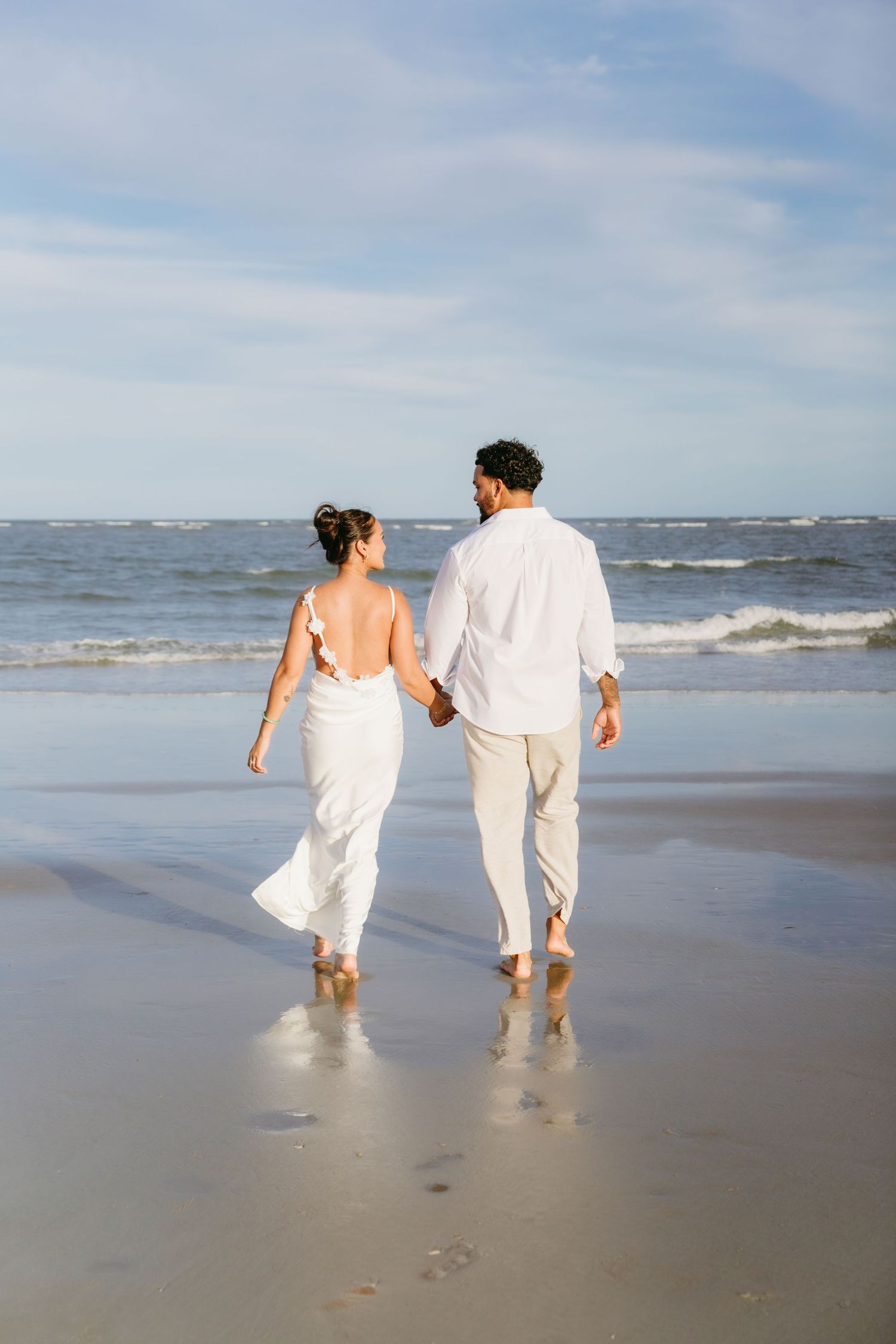 Couple in white walking hand-in-hand along shoreline with ocean waves behind them.