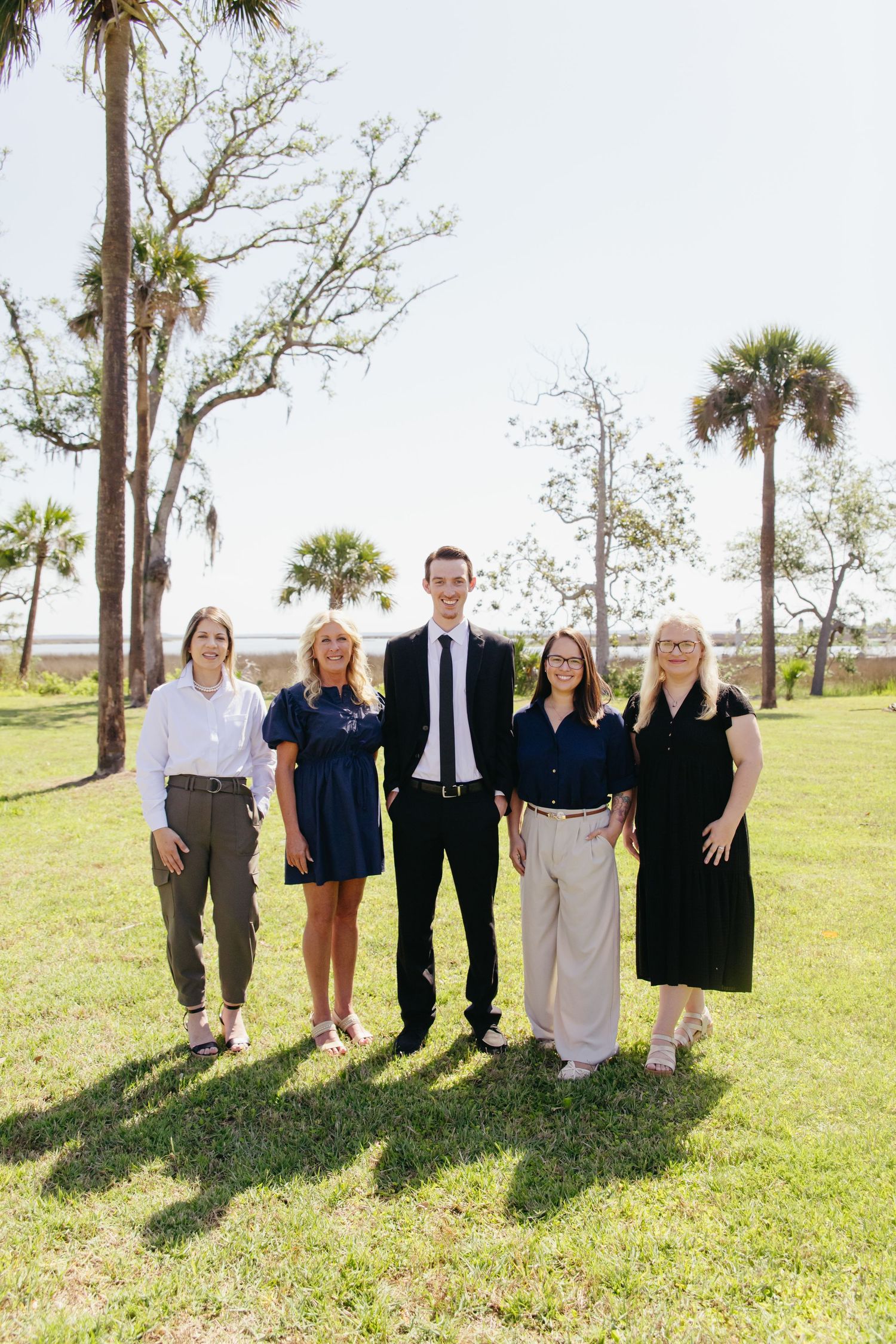 Group of five professionally dressed colleagues posing together on green lawn with palm trees and water behind them.