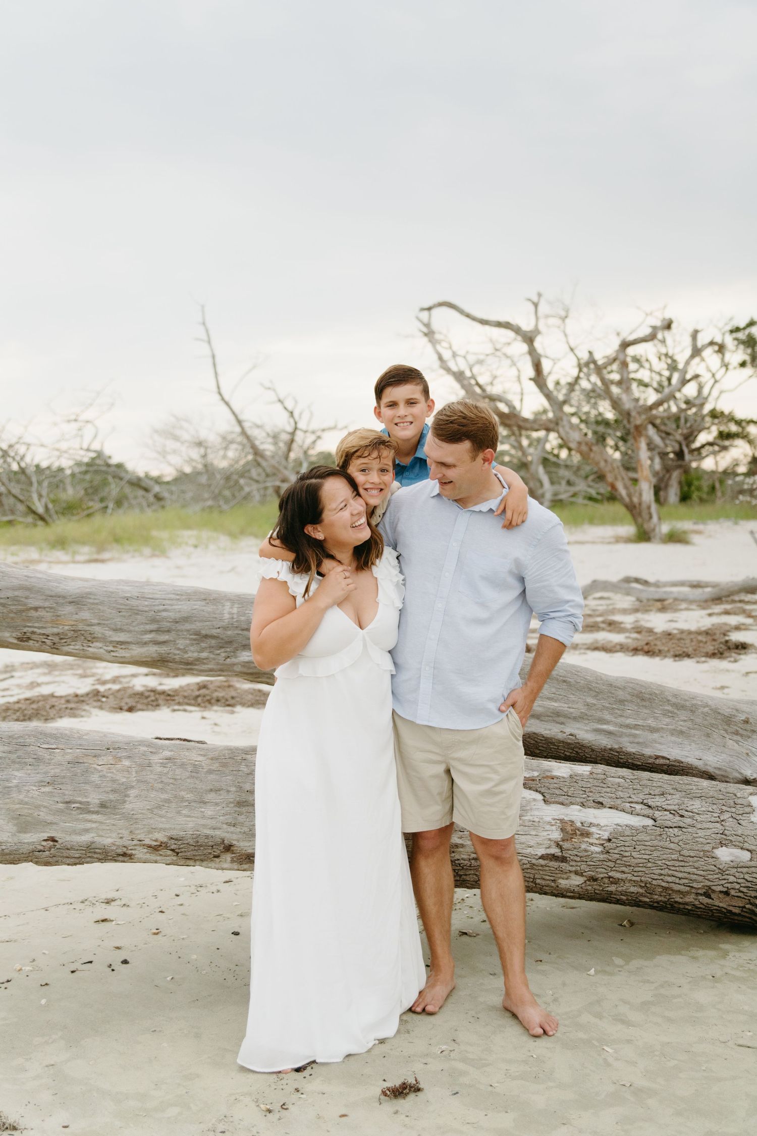 Happy family of four posing together on a sandy beach with driftwood and trees behind them.