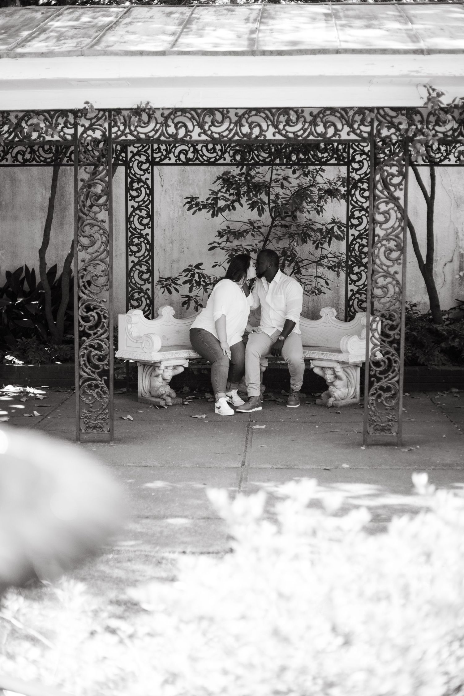 Black and white photo of couple sharing a kiss on a bench beneath an ornate iron gazebo.