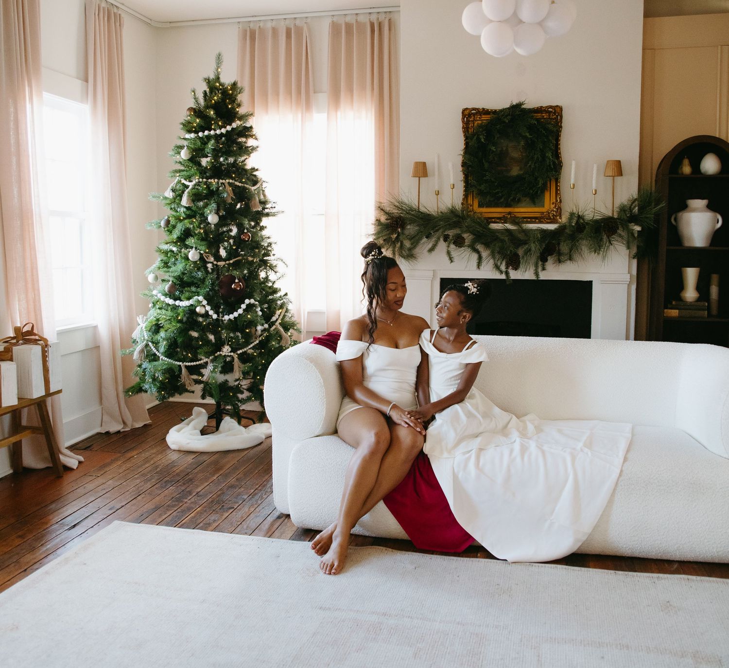 A mother and daughter in white dresses relax on a white sofa beside a decorated Christmas tree in an elegant living room.
