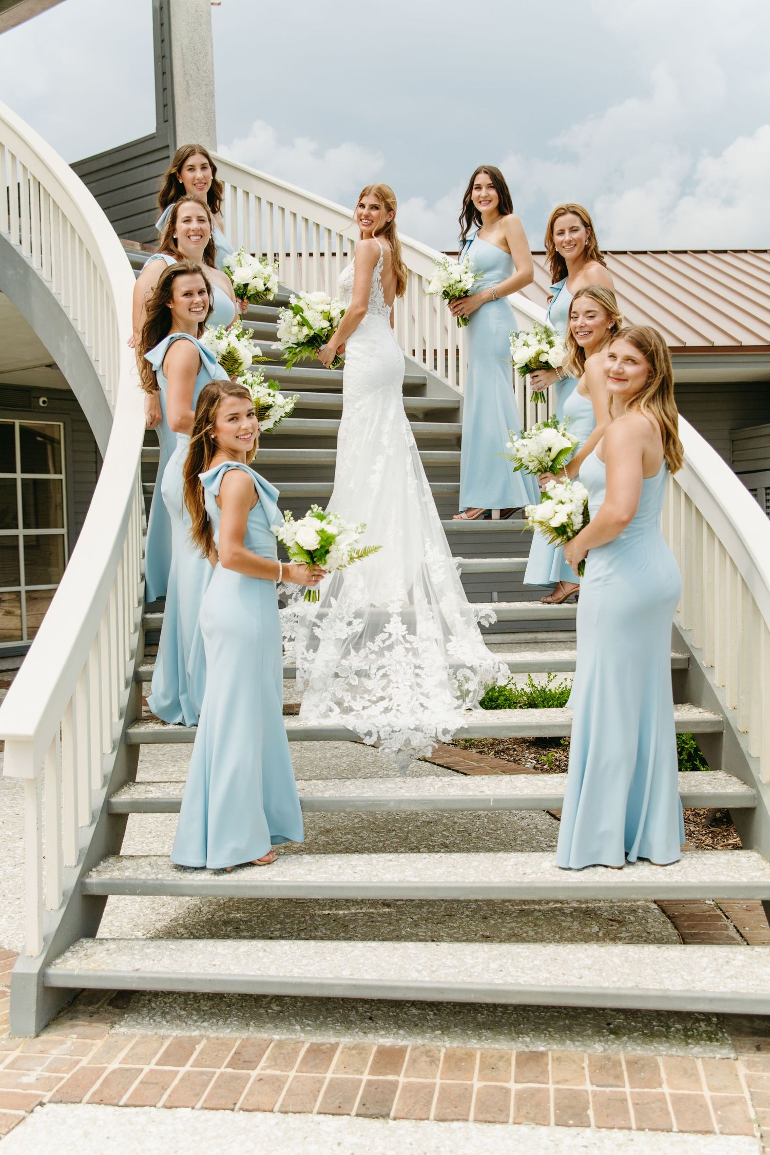 Bride in lace gown poses with bridesmaids in light blue dresses holding white bouquets on elegant staircase.