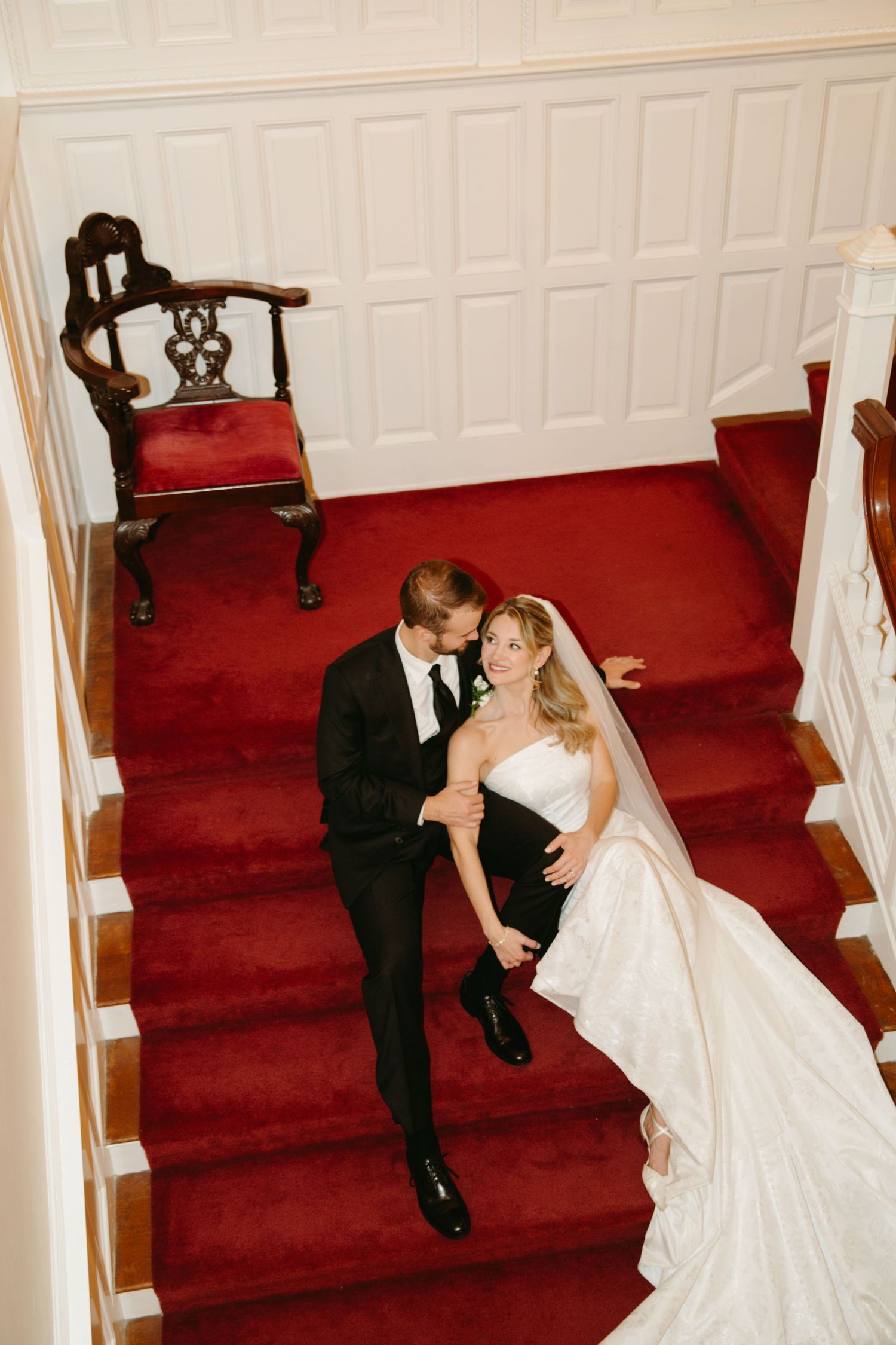 Bride and groom pose romantically on grand staircase with red carpet inside elegant wedding venue.