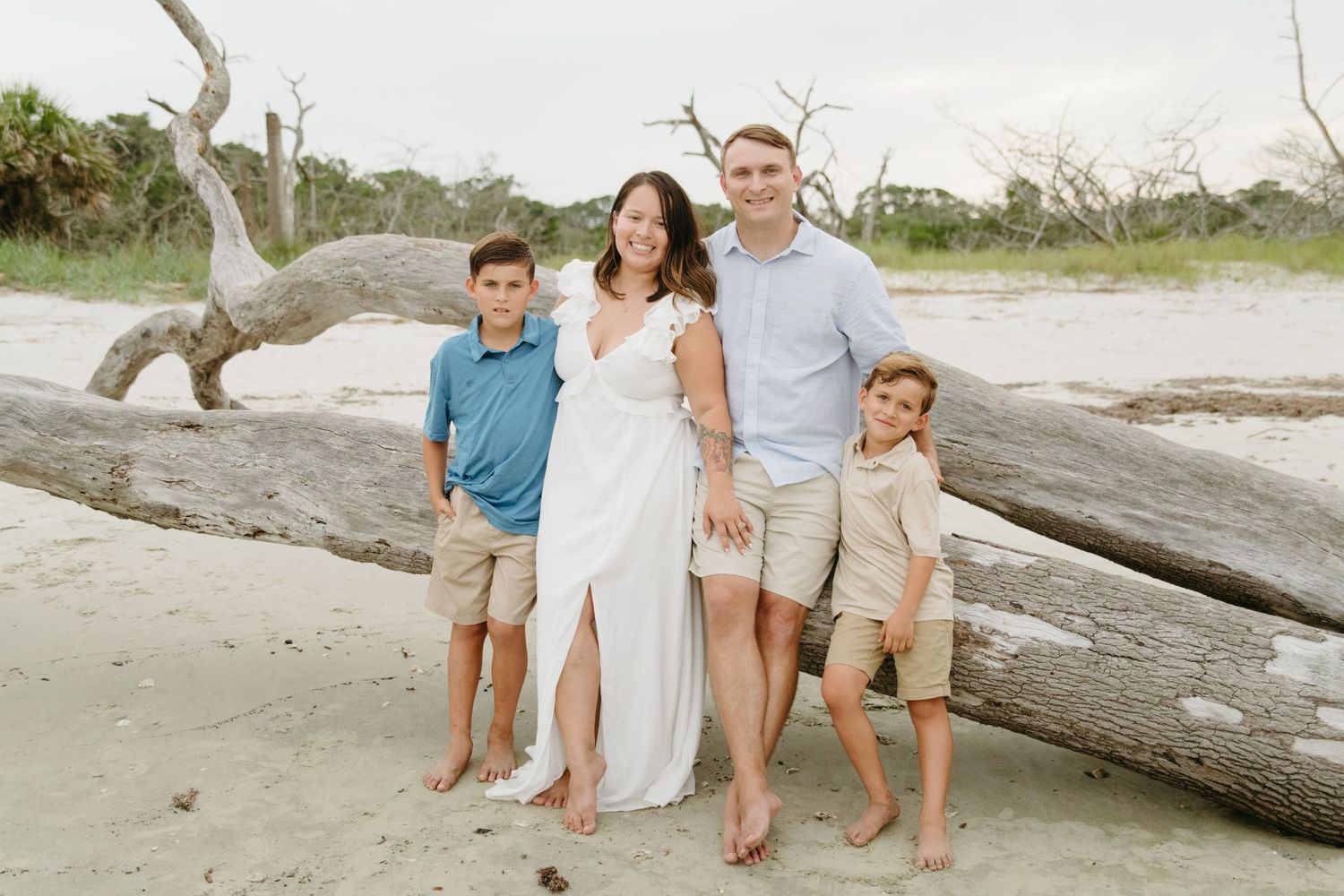Happy family of four posing by driftwood on a sandy beach on a cloudy day.