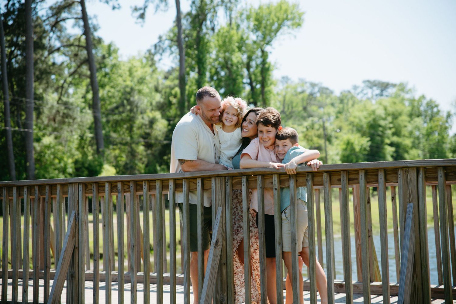 Family of five leans on wooden bridge railing surrounded by lush green trees.