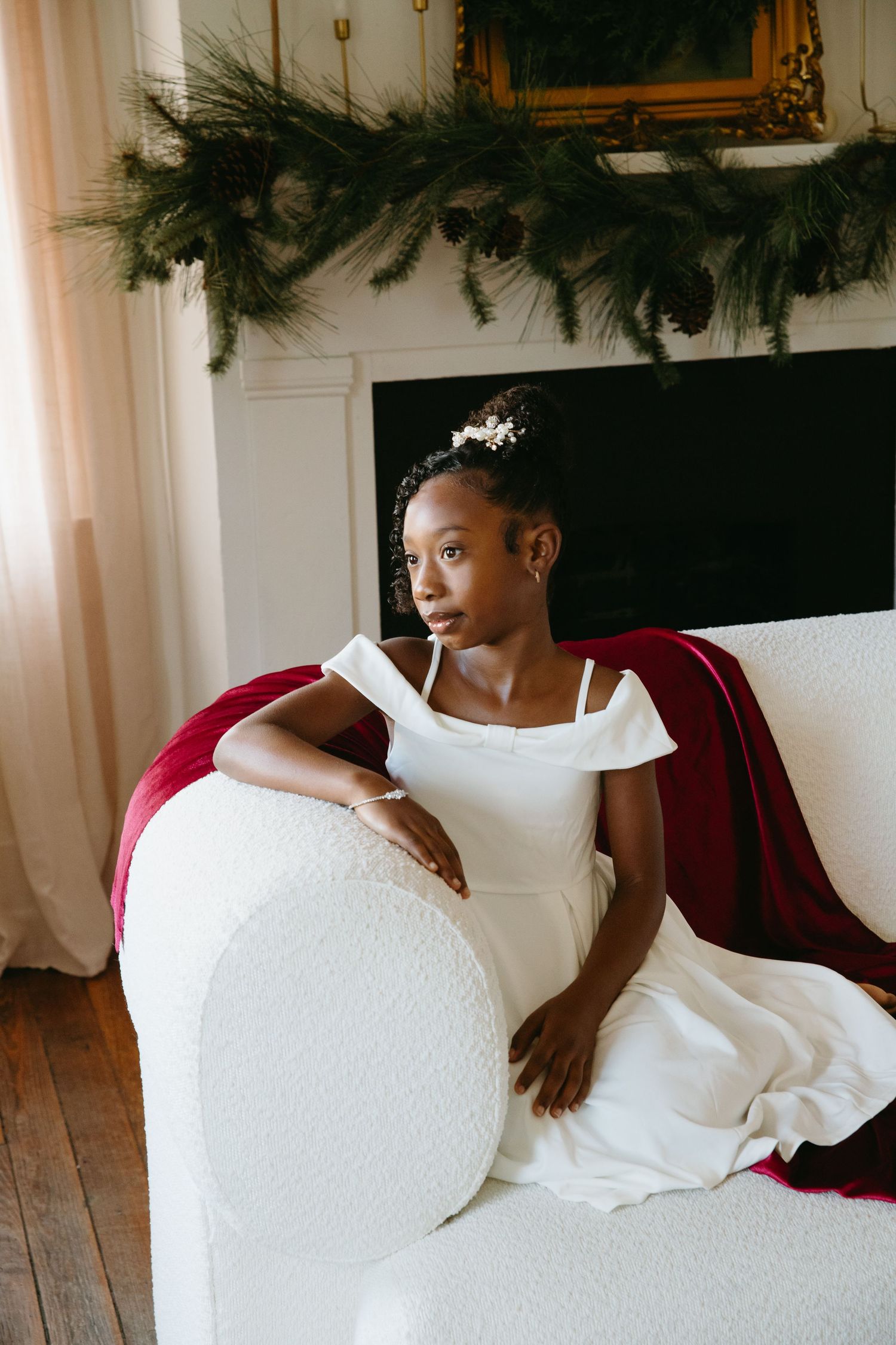 A young girl in a white dress with floral hair accessory reclines gracefully on a white sofa near a garland-adorned fireplace.