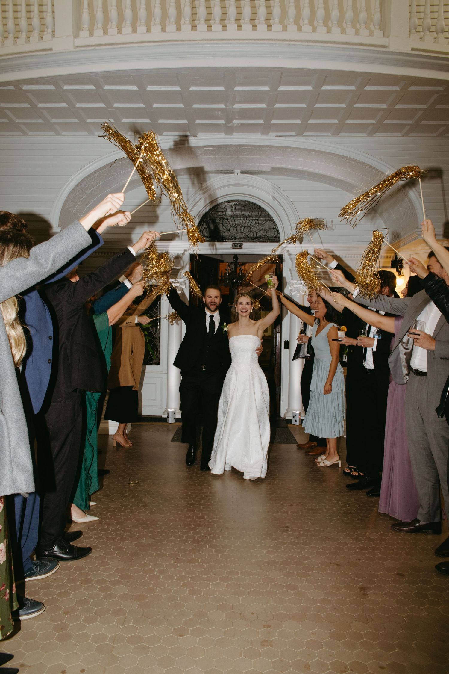 Bride and groom exit wedding reception through sparkler tunnel with cheering guests lining the hallway.