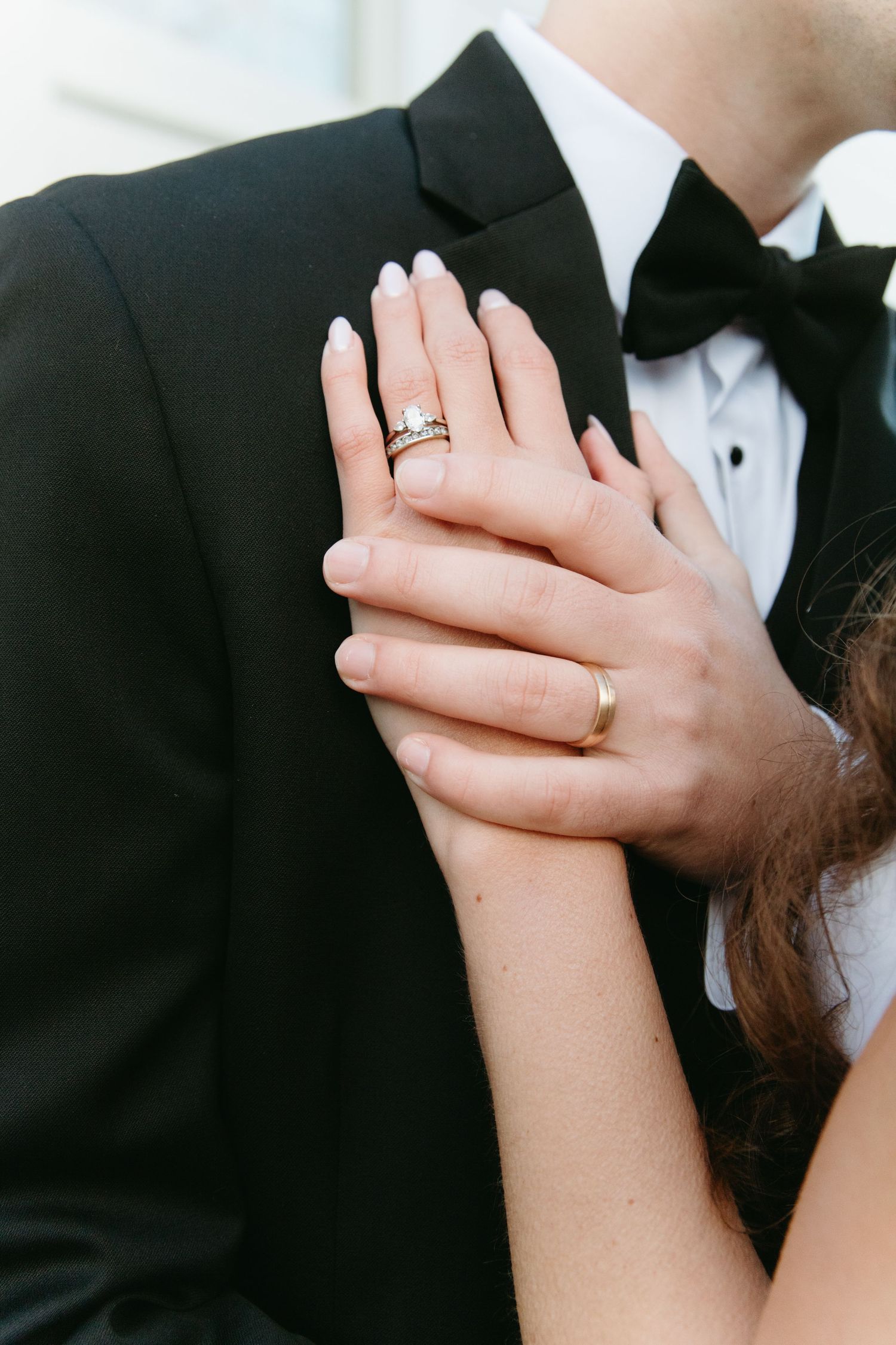 Close-up of bride and grooms hands showing wedding rings against black tuxedo.