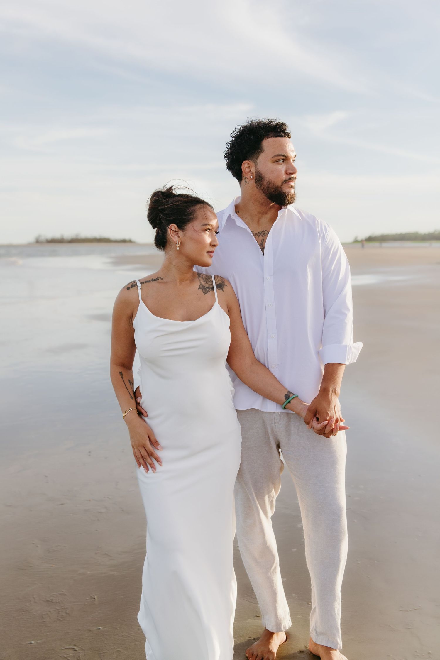 Couple in white outfits holding hands on a beach, woman in slip dress and man in linen shirt.