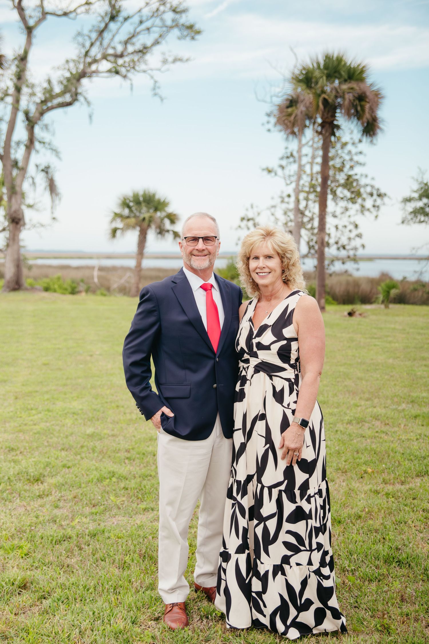 Smiling couple posing outdoors on green lawn, man in navy blazer with red tie, woman in black floral maxi dress.