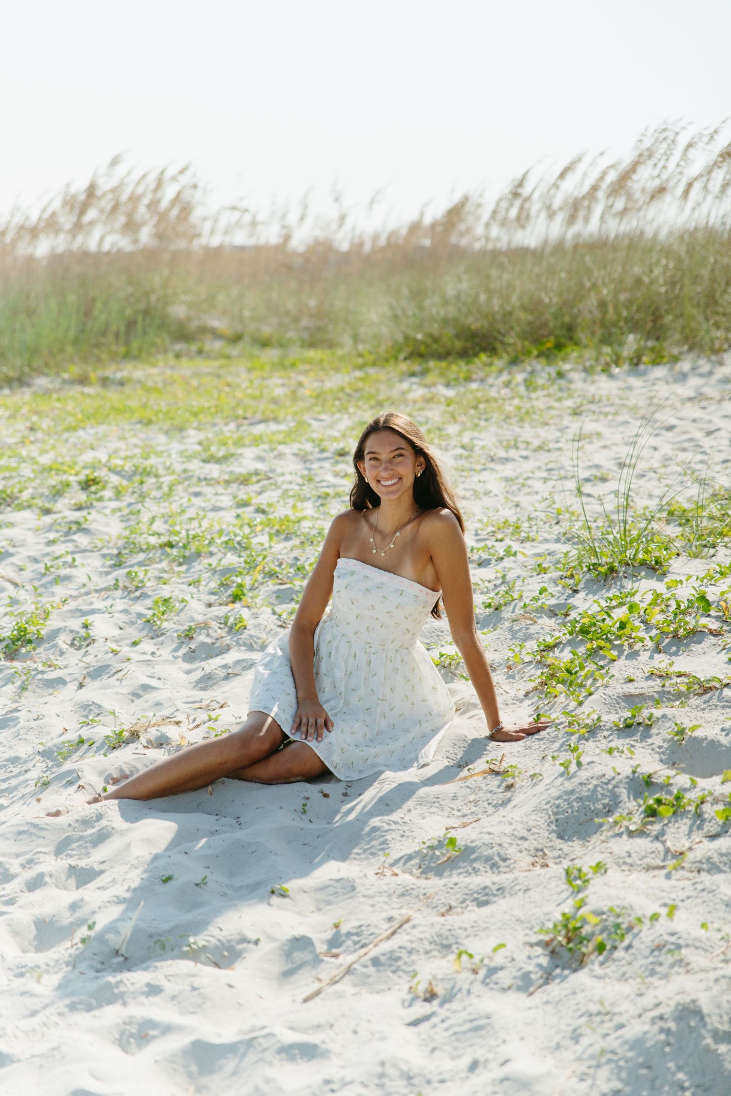 Smiling young woman in white sundress sitting on white sand beach dunes on a sunny day.
