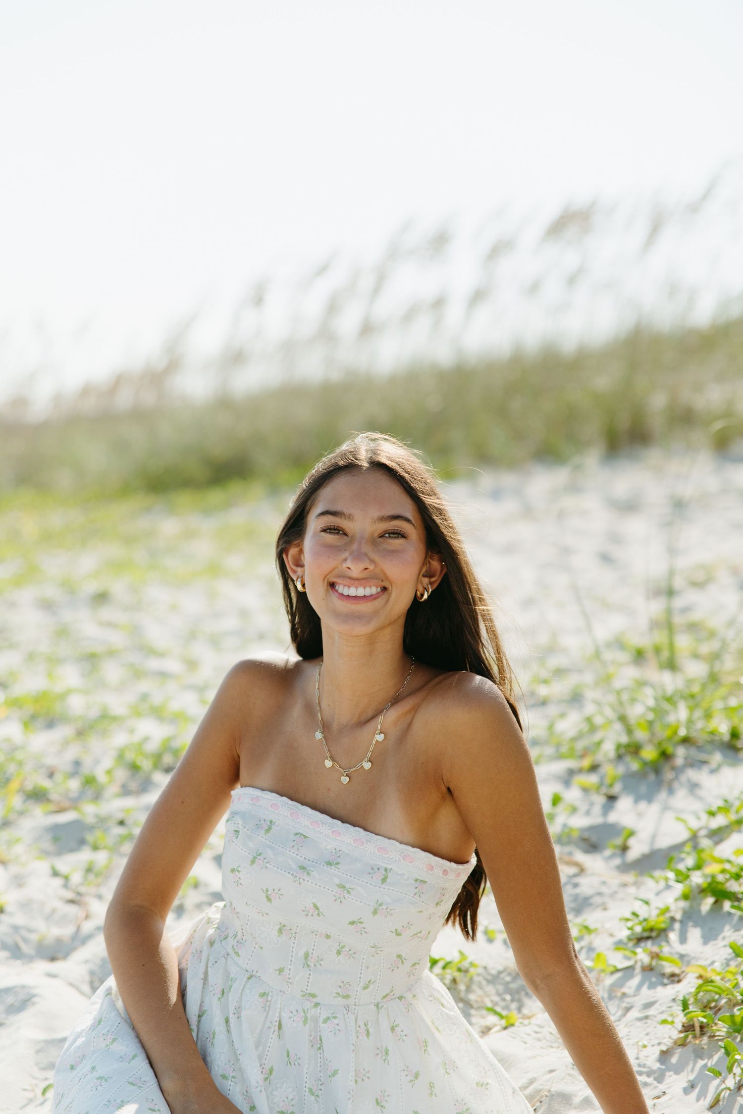 Smiling young woman in a floral strapless dress sitting on sandy beach dunes with sea oats.