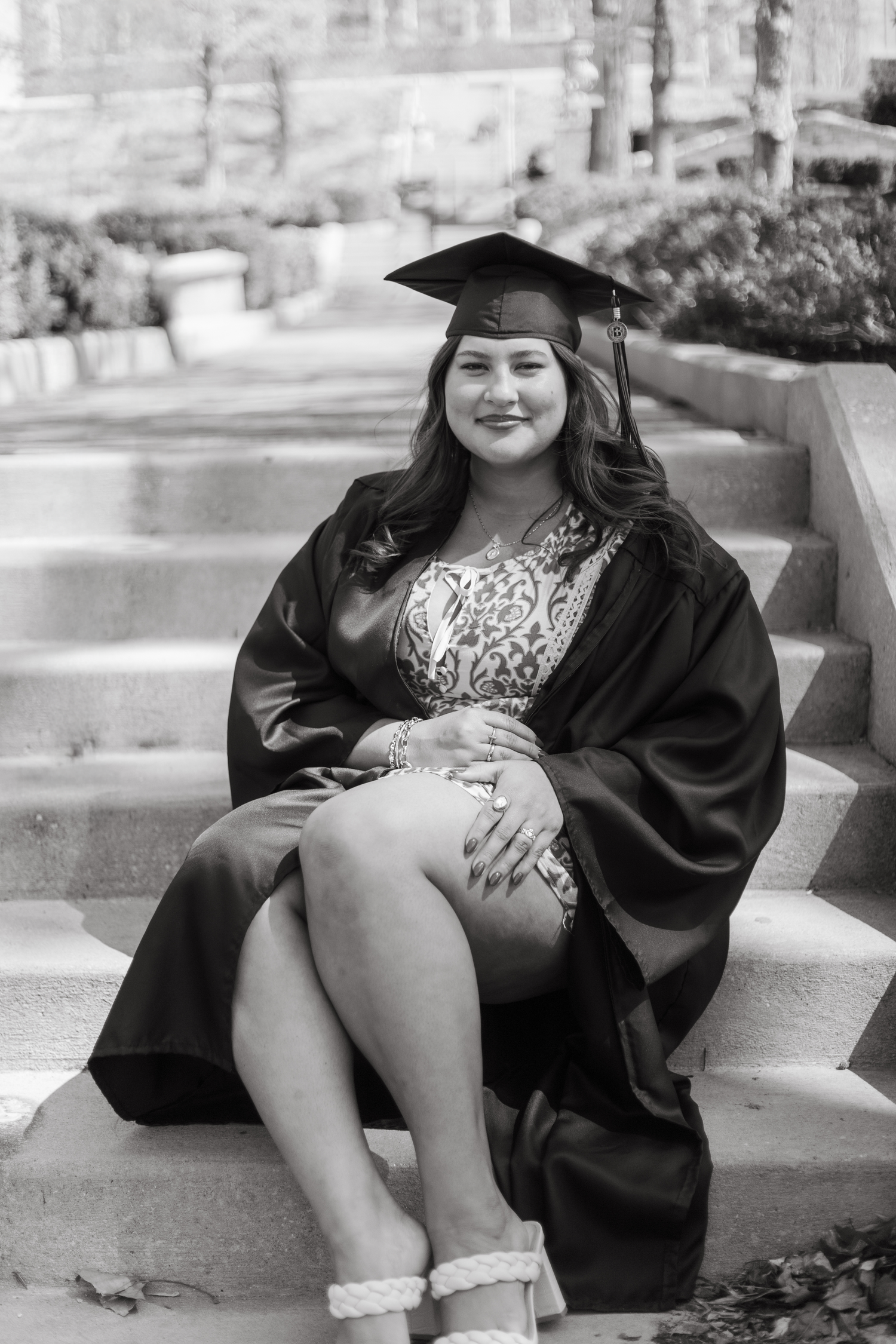 Black and white graduation portrait of a woman in cap and gown sitting on stone steps outdoors.