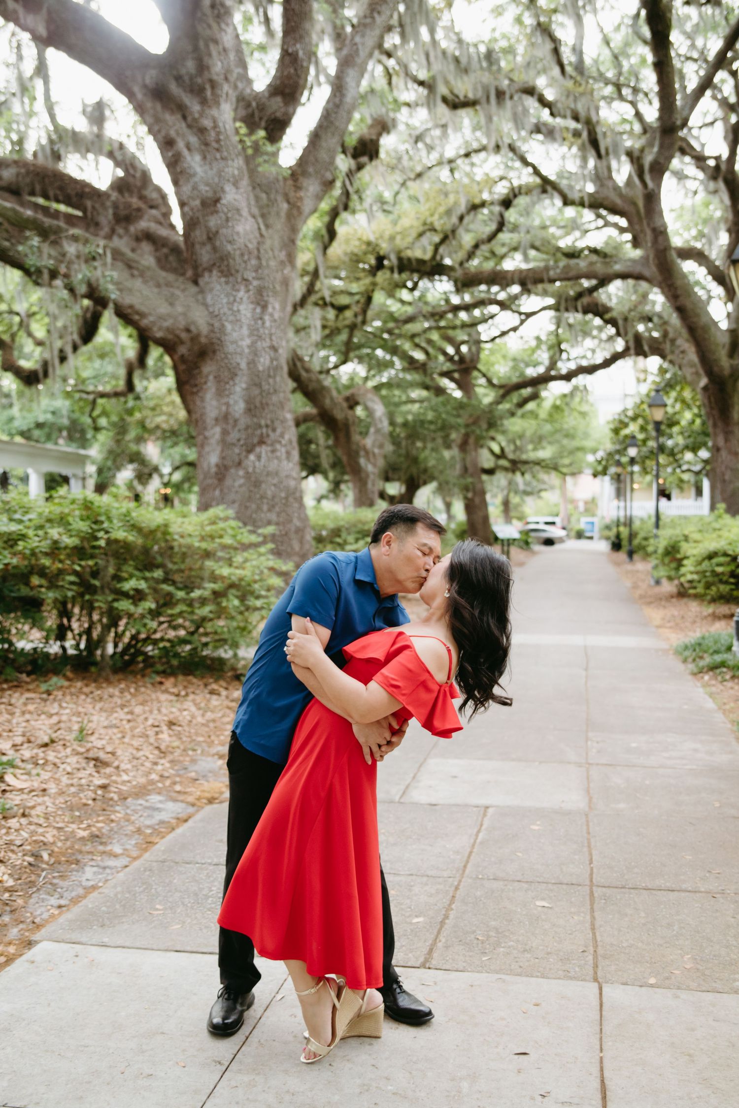 Couple sharing a romantic dip kiss on a scenic sidewalk lined with large oak trees.