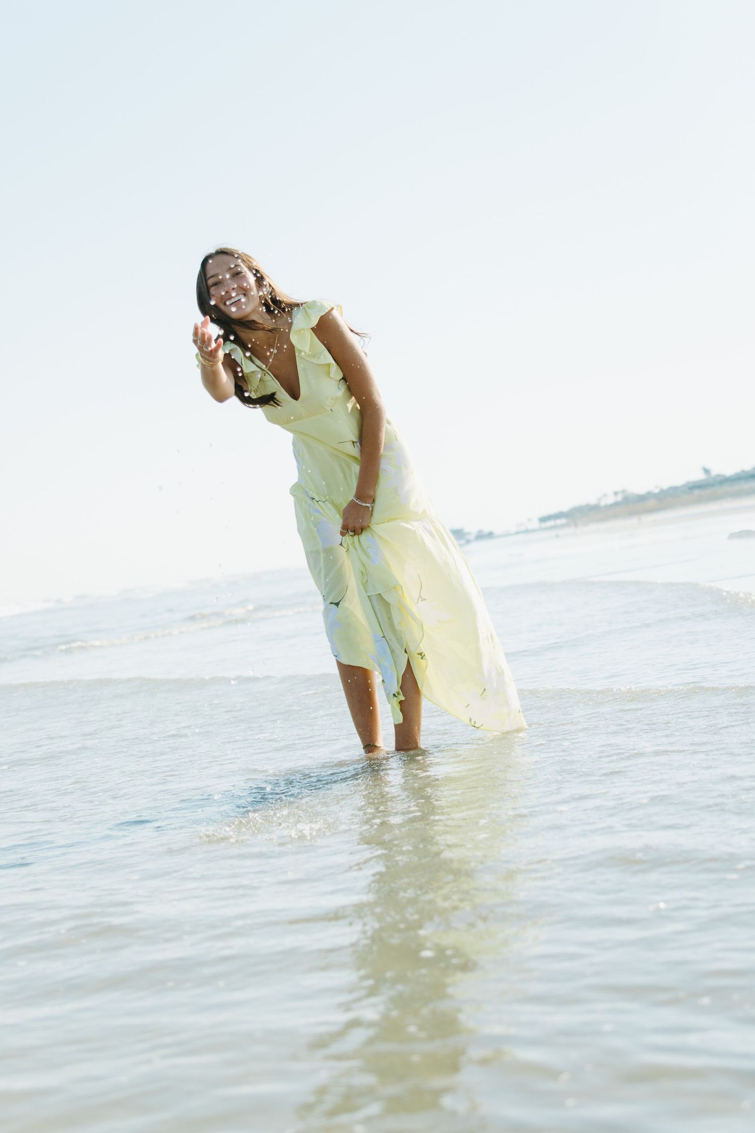 Joyful woman in a yellow dress standing in ocean waves at a sunny beach, holding a seashell.