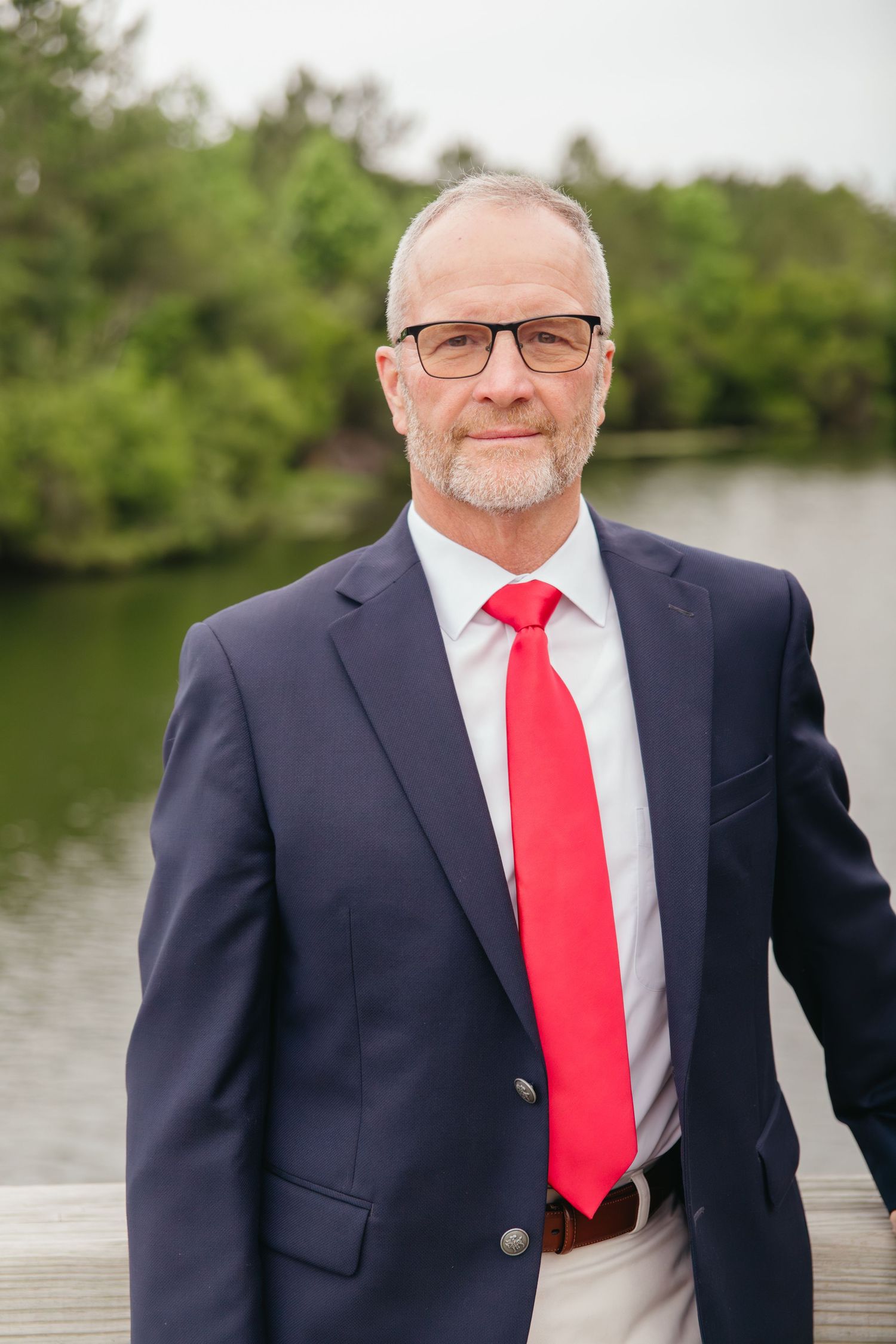 Distinguished older man in navy suit with red tie wearing glasses posing near waterfront with green trees behind him.