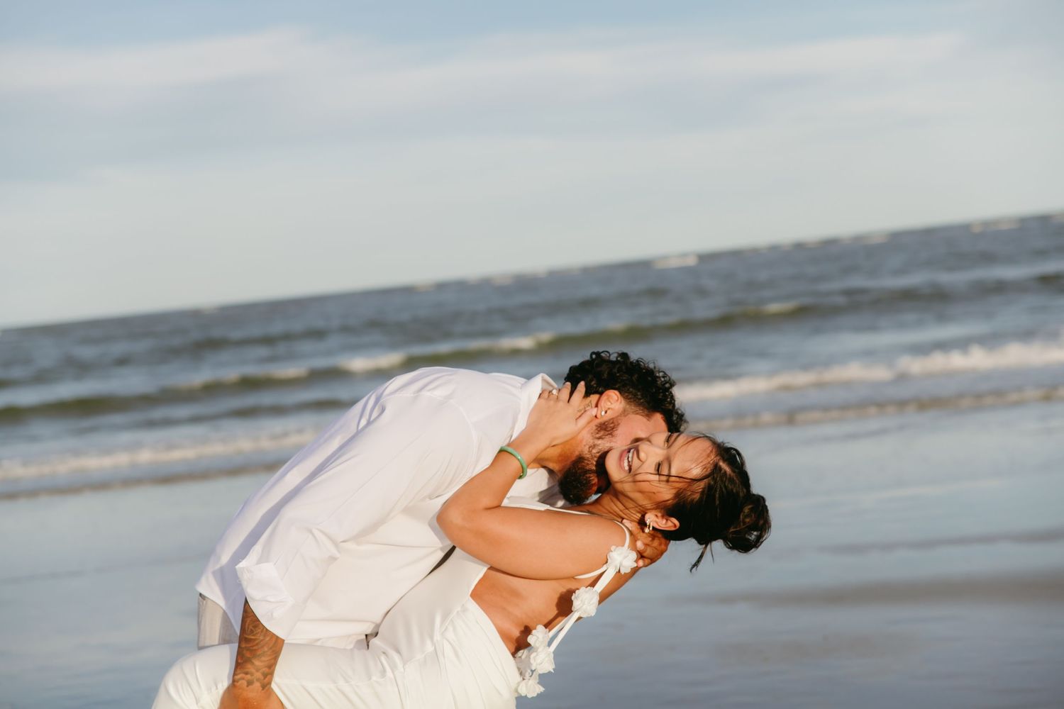 Joyful couple laughing and embracing on a beach with ocean waves in the background.