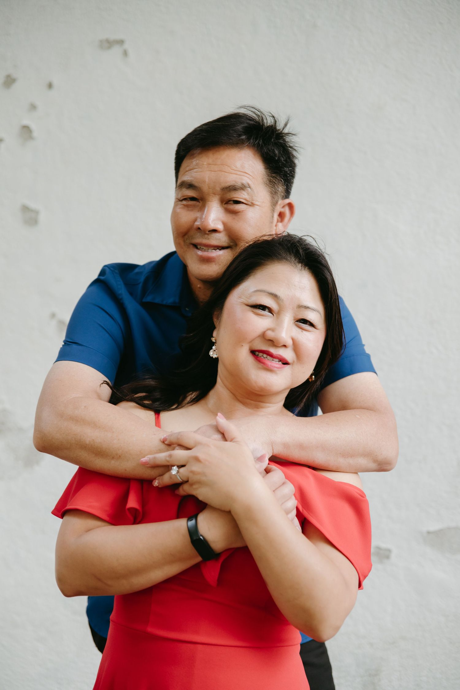Smiling Asian couple embracing warmly against a white wall, man hugging woman from behind.
